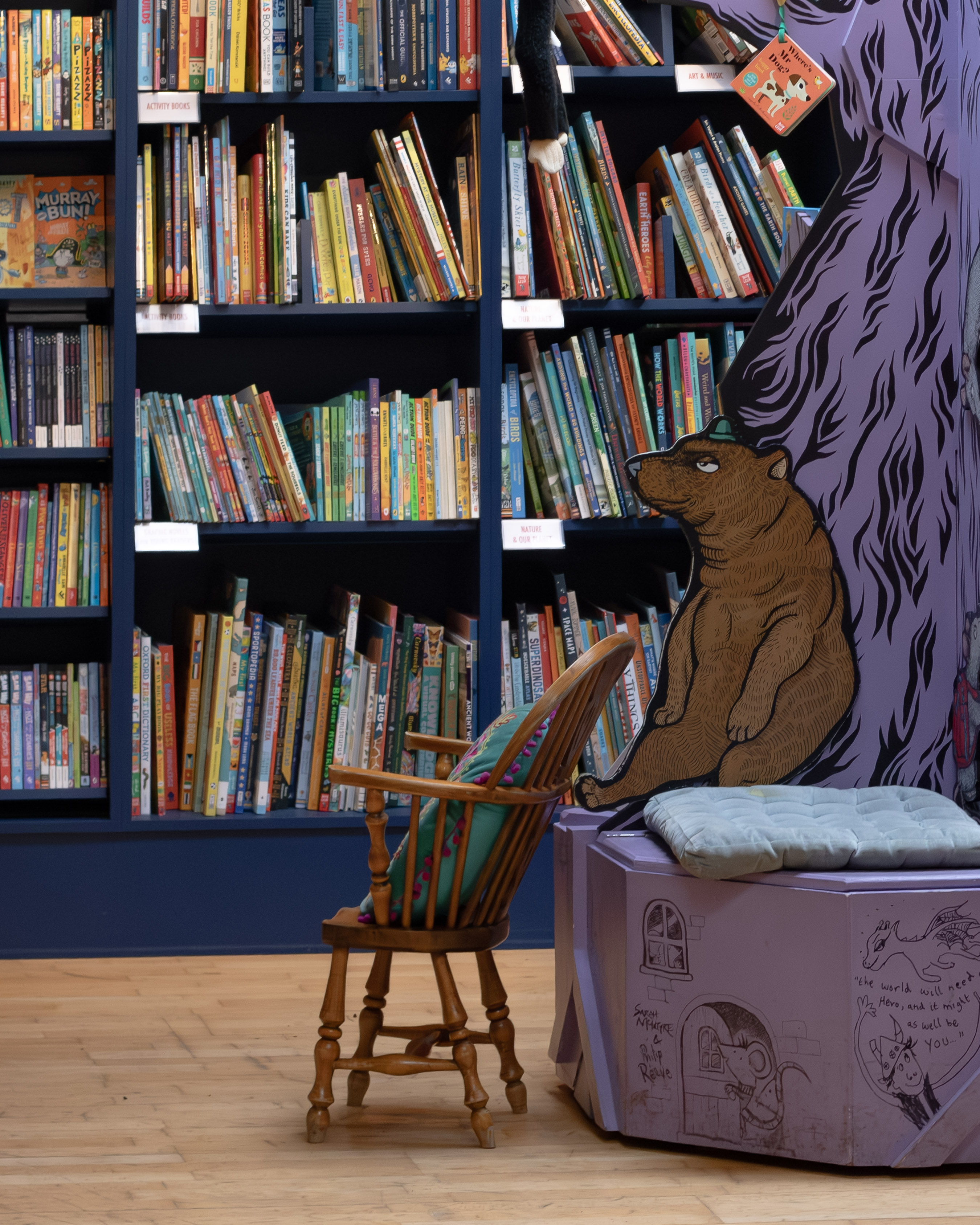 Children's section of a bookshop with a large painted bear illustration and an empty wooden chair beside bright bookshelves
