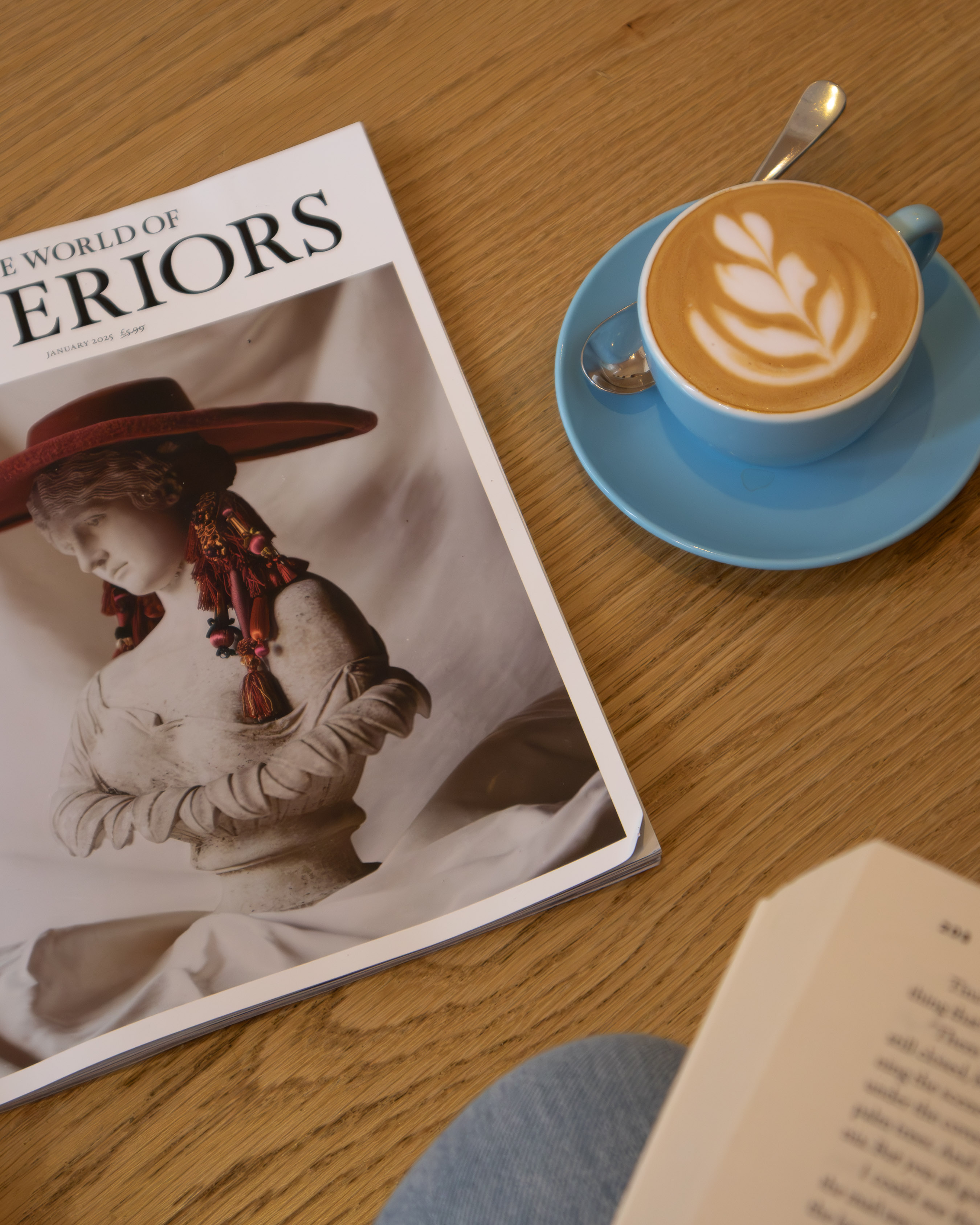 Copy of The World of Interiors magazine beside a blue cup of latte-art coffee on an oak cafe table