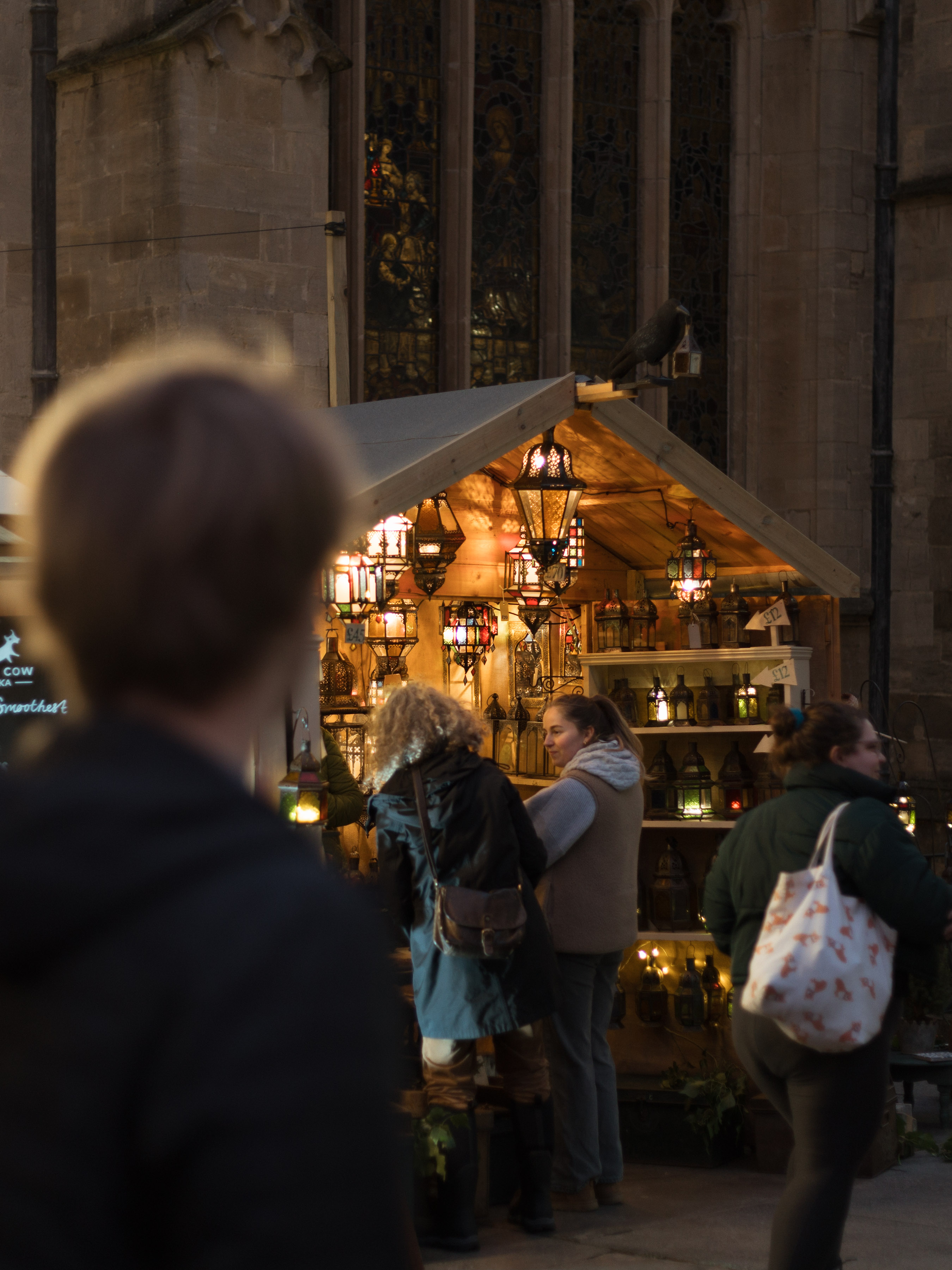 Lantern-lit wooden chalet at the Bath Christmas Market in front of stained glass of the Abbey at dusk
