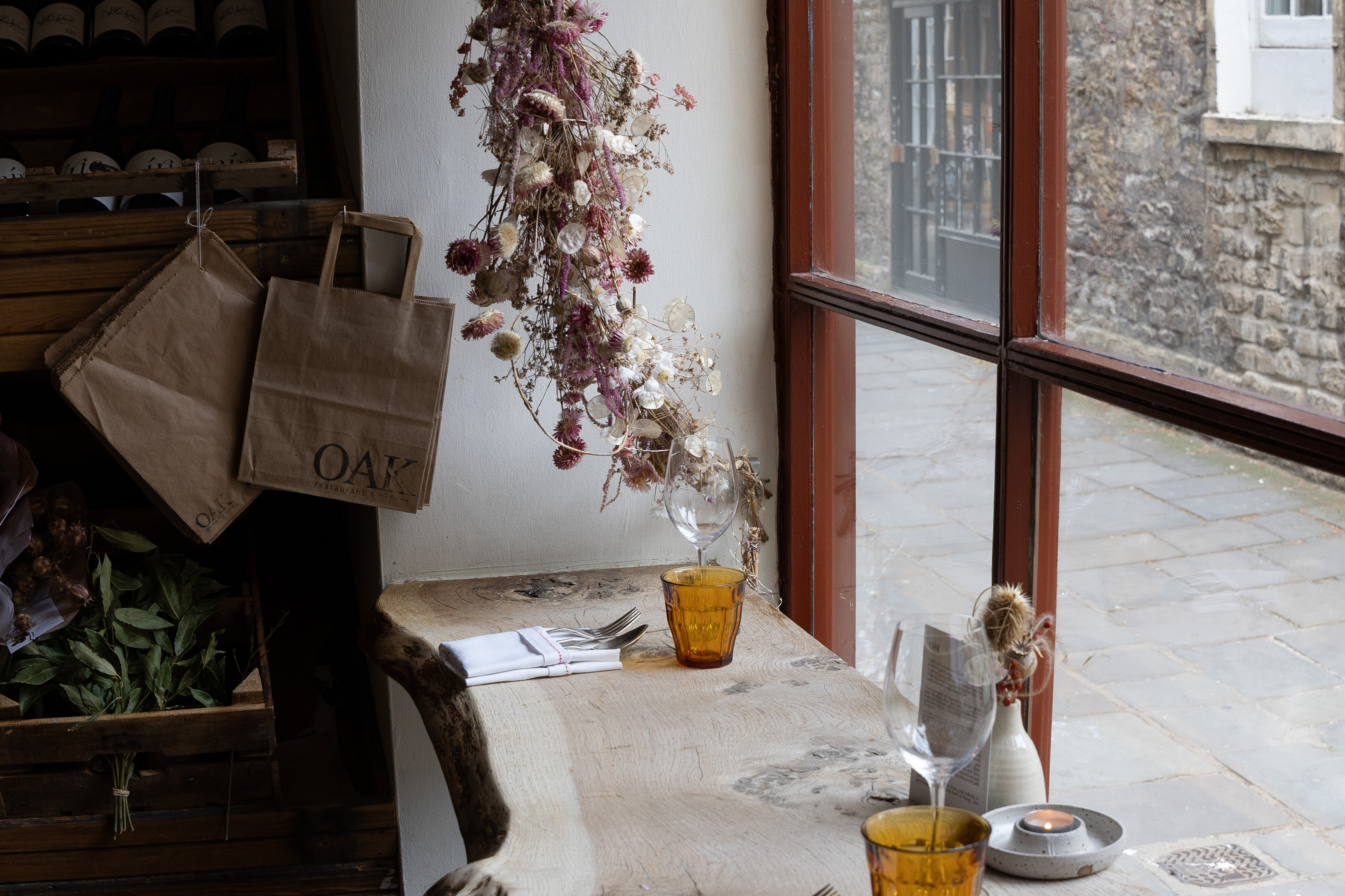Sunlit cafe corner with paper grocery bags, dried flowers and amber-glass tumblers on a rustic timber bench