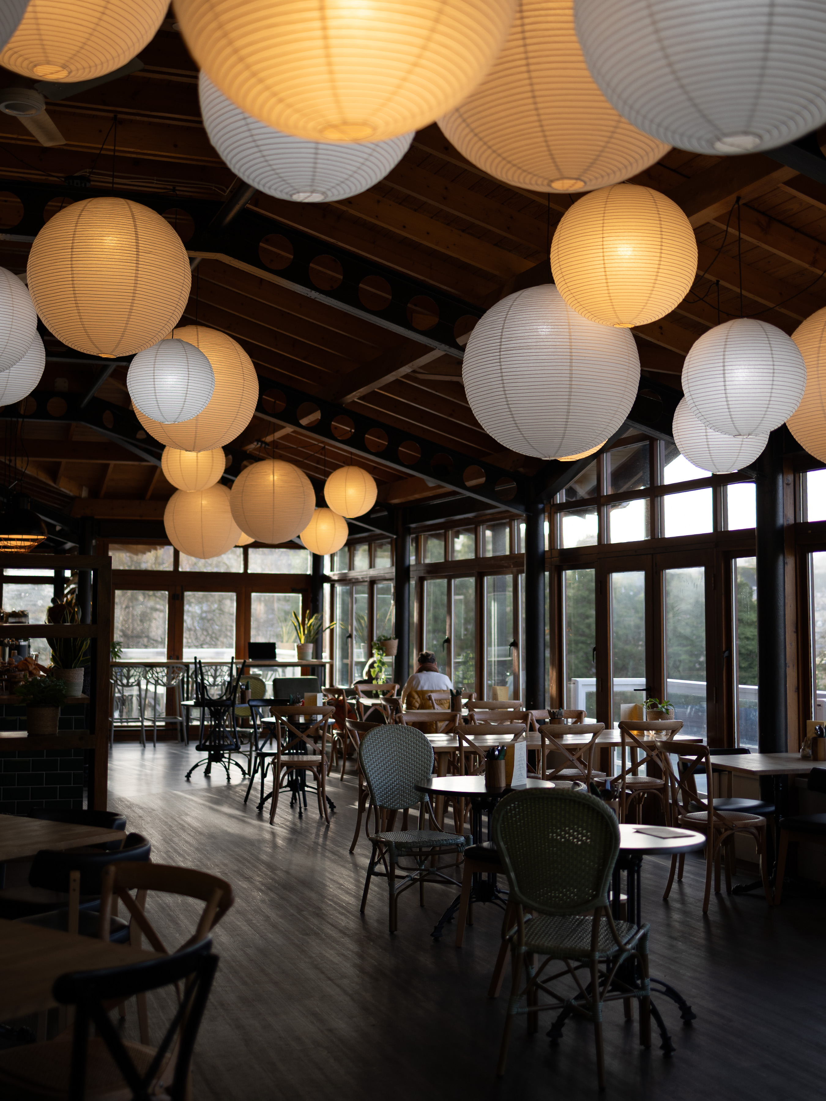 Restaurant interior with cascading paper lantern lights above bistro tables and large windows looking onto woodland
