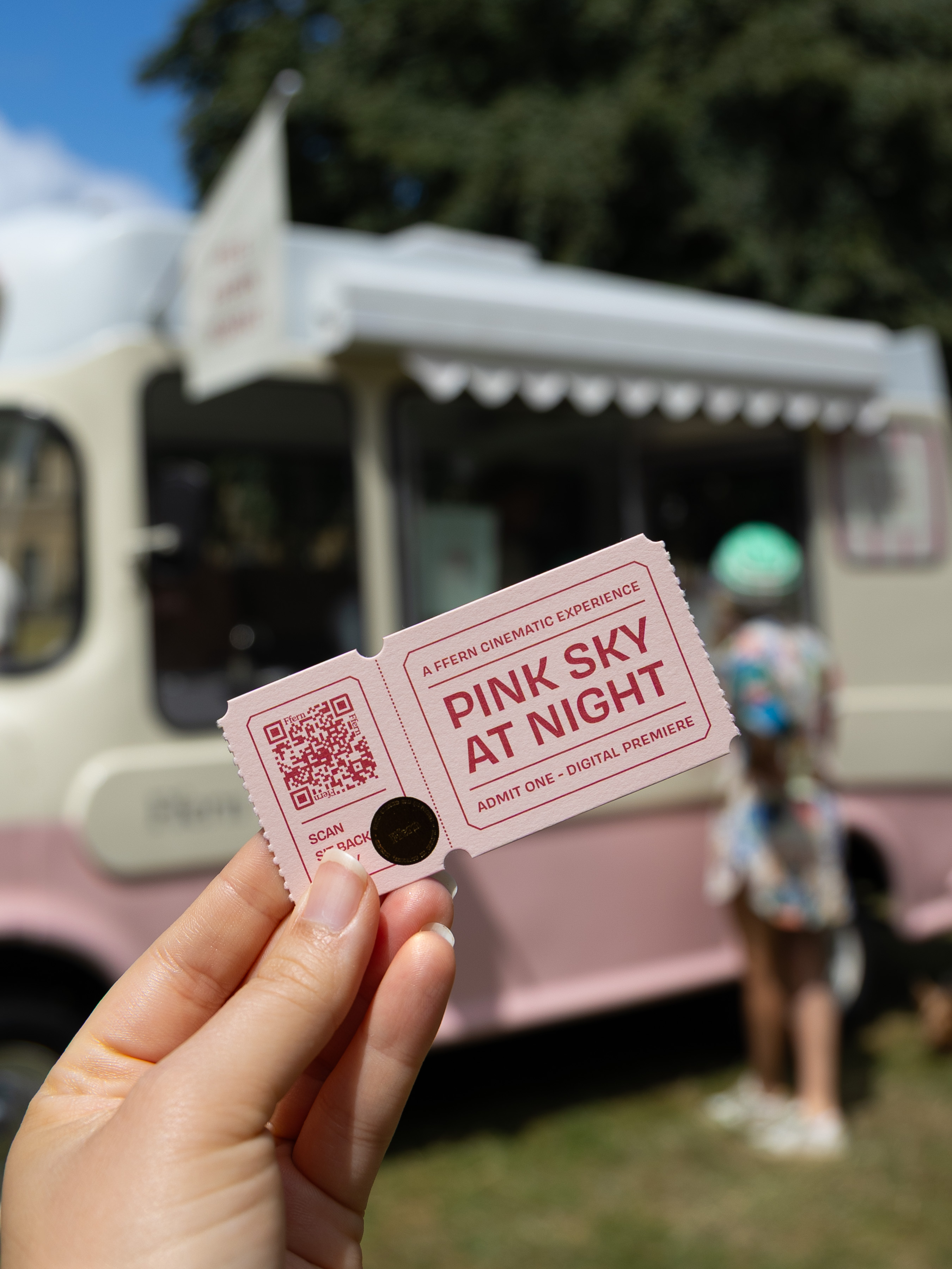 Hand holding a pink admission ticket in front of a vintage ice-cream van at a summer event
