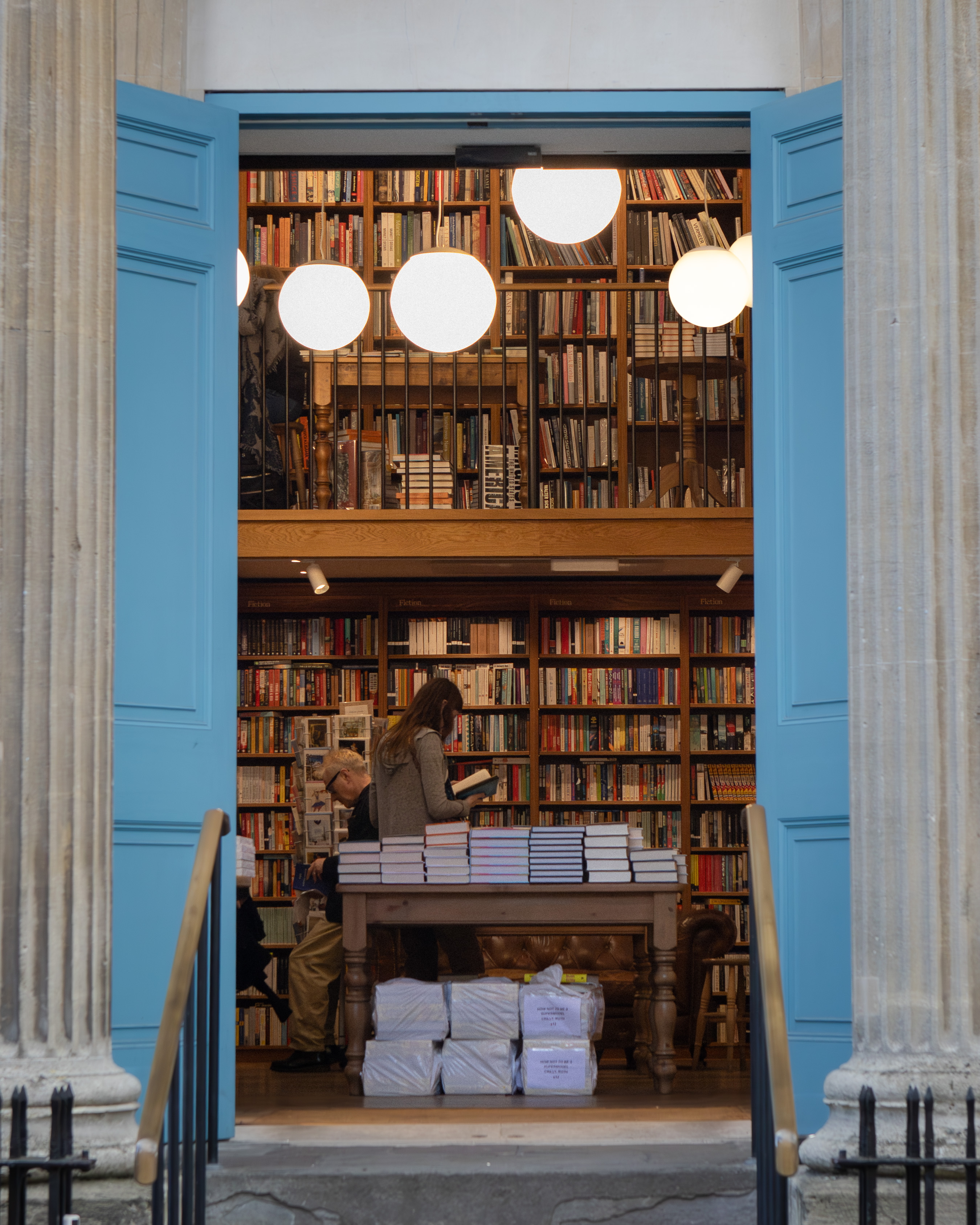 Customers inside a classical-portico bookshop browsing tables of books beneath spherical pendant lights