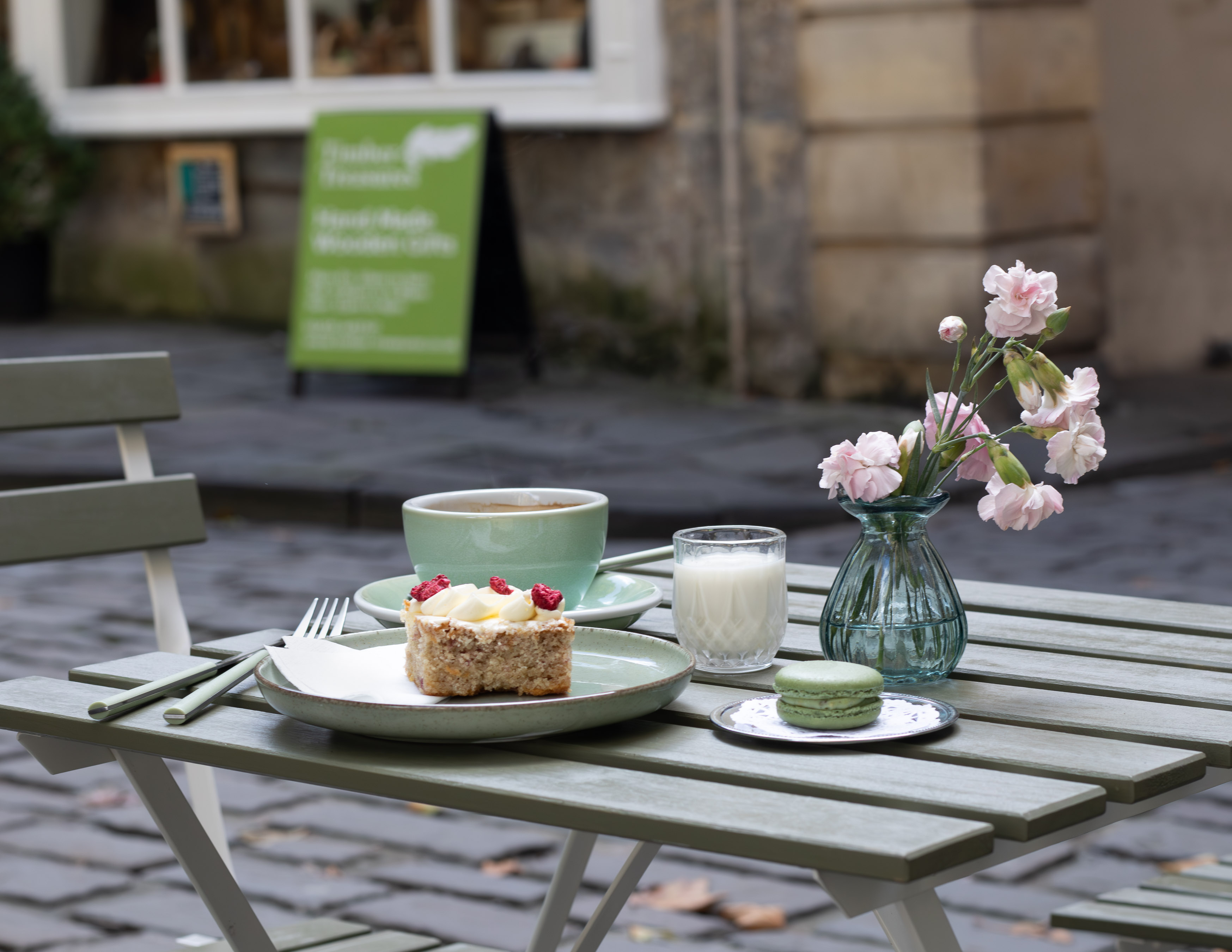 Cafe terrace table with cappuccino, sponge cake and pink carnations in front of a Bath cobblestone alley