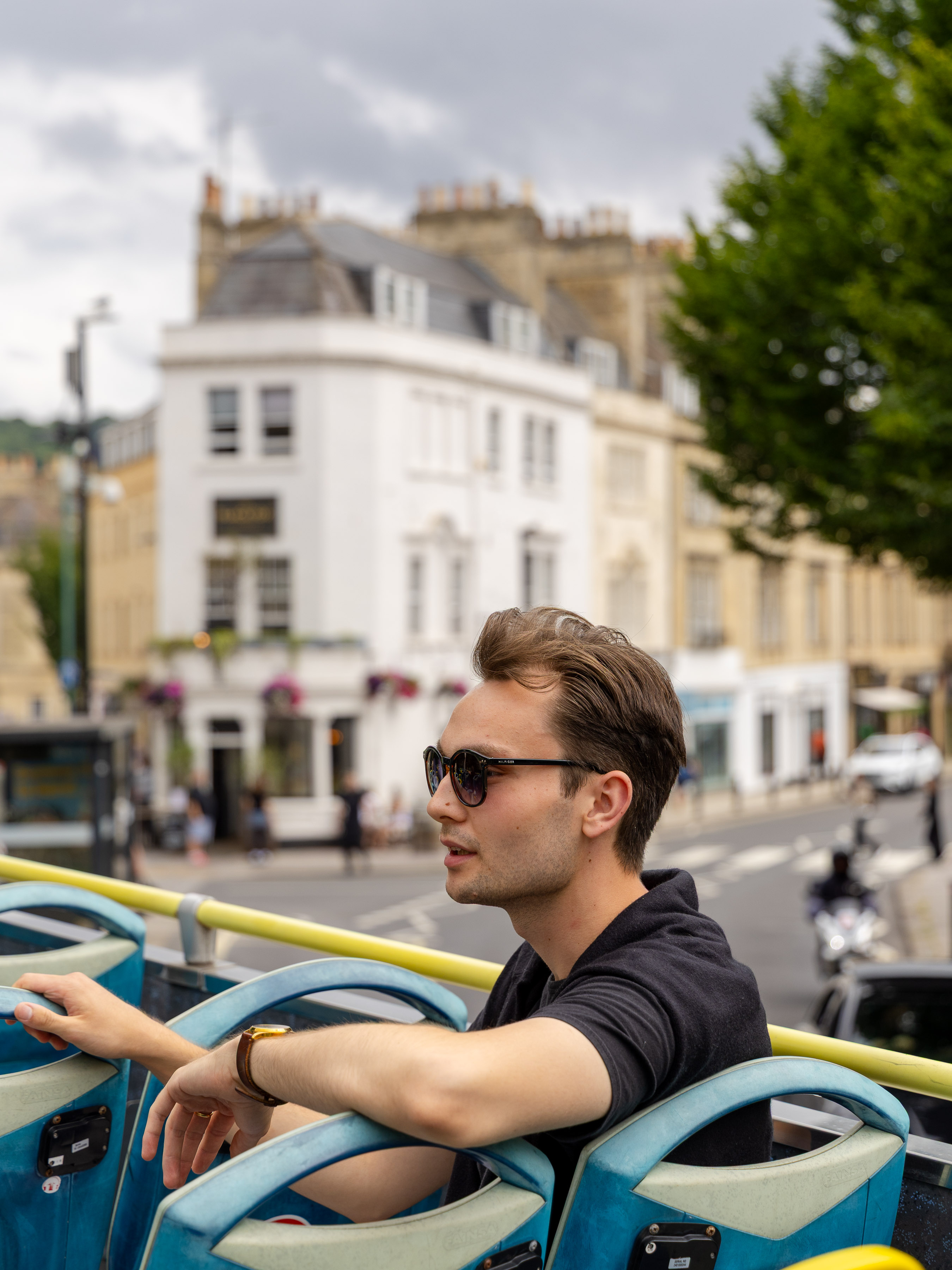 Young man in sunglasses on the upper deck of an open-top sightseeing bus passing through Bath
