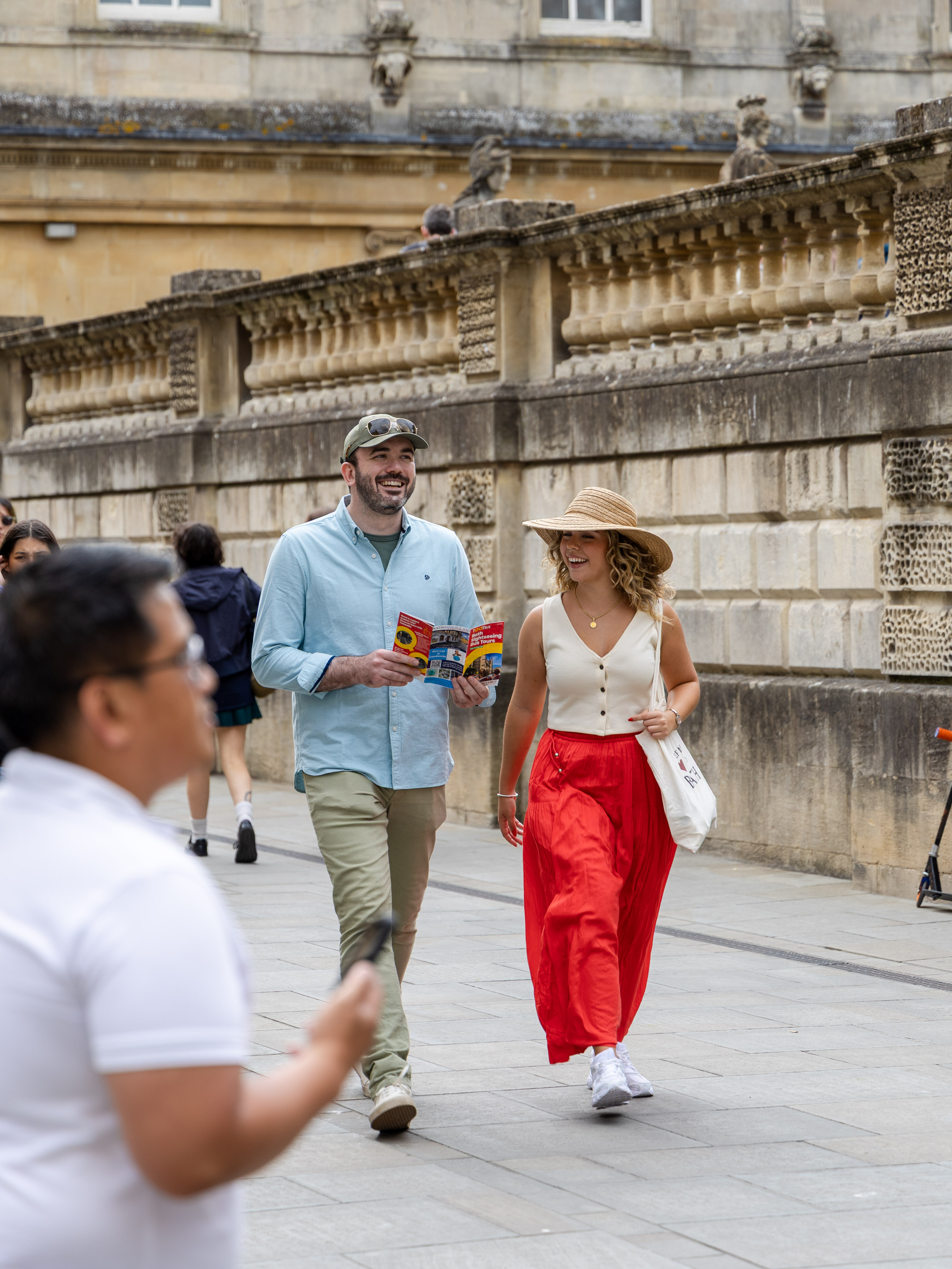 Two visitors with a Bath tourist map walking past the Roman Baths, with guide in foreground