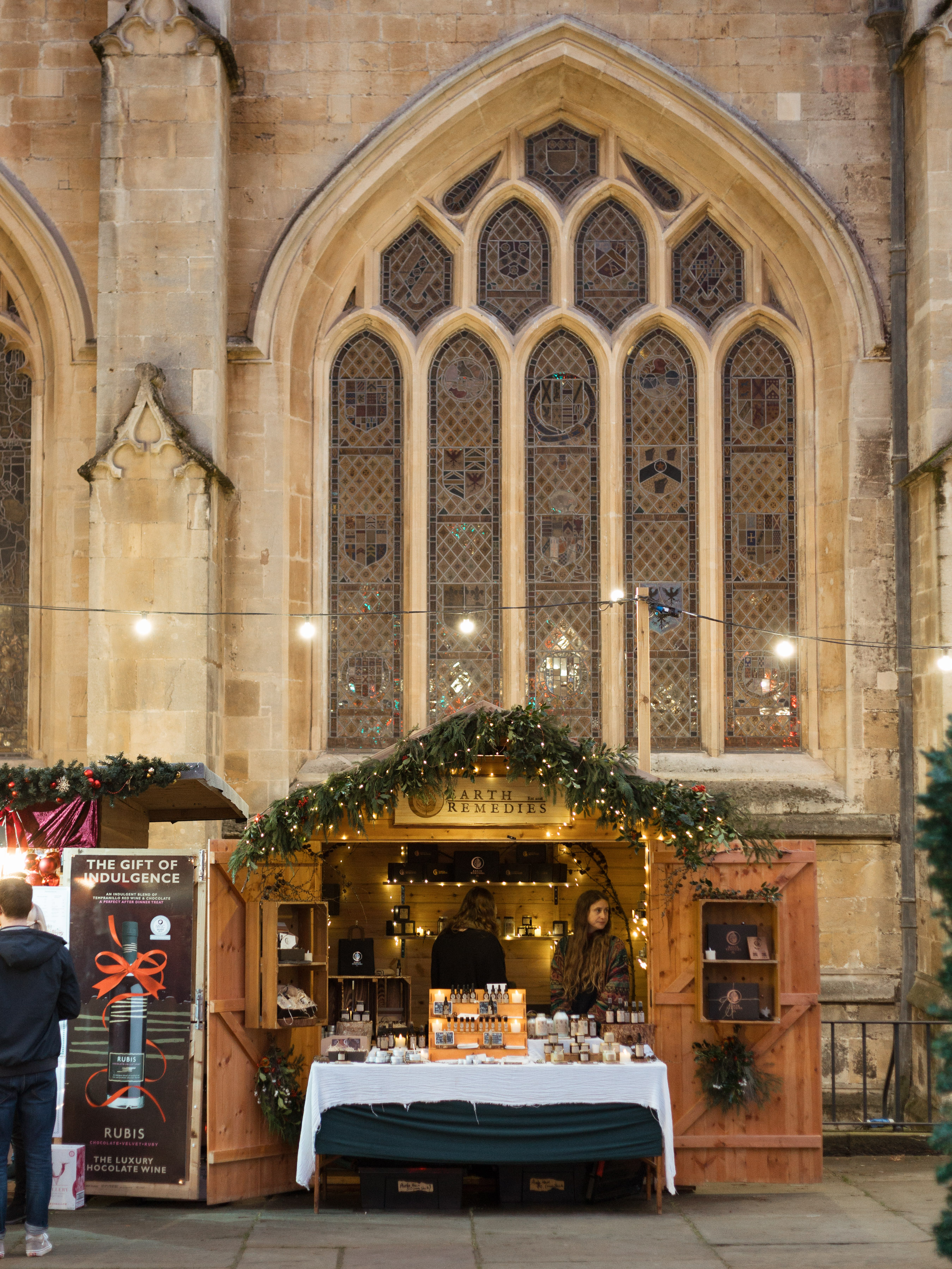 Bath Christmas Market chalet stall stocked with apothecary bottles, set against the stained-glass windows of Bath Abbey