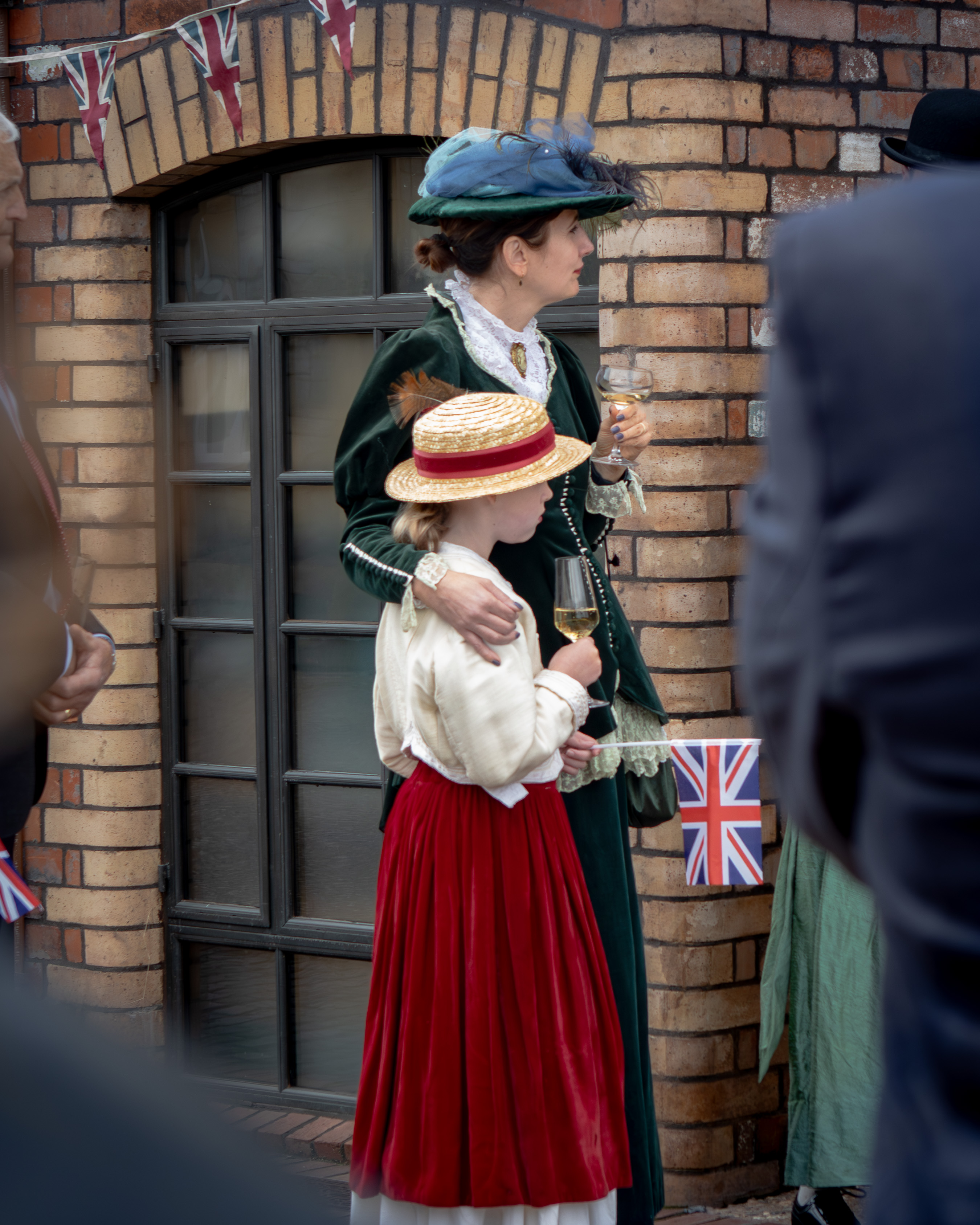 Regency-costumed mother and daughter in green velvet and red skirts holding wine glasses beneath bunting at a celebration