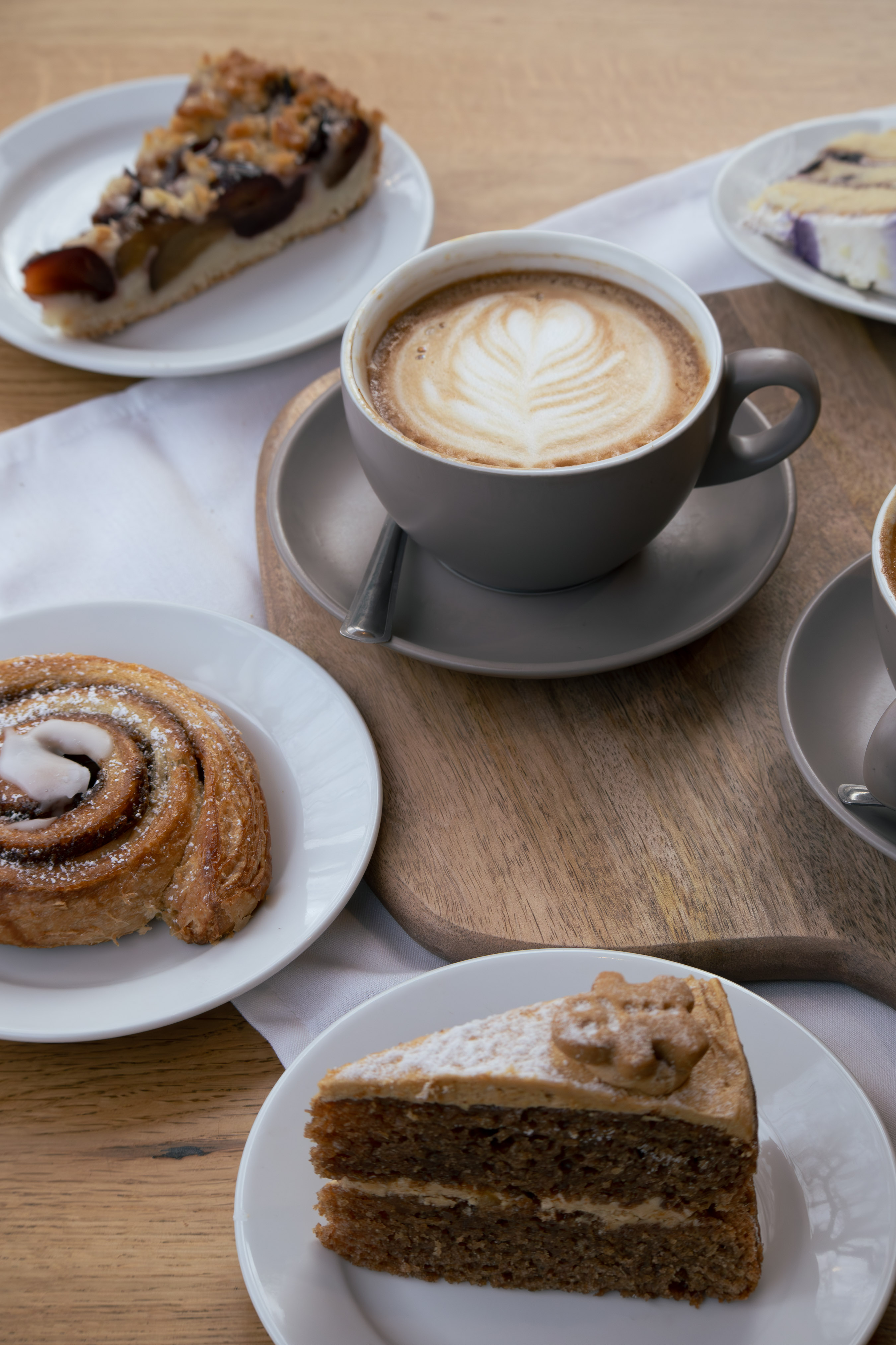 Flat-lay of cafe pastries: cinnamon swirl, plum tart, blueberry cheesecake and a coffee cake around a latte-art cappuccino