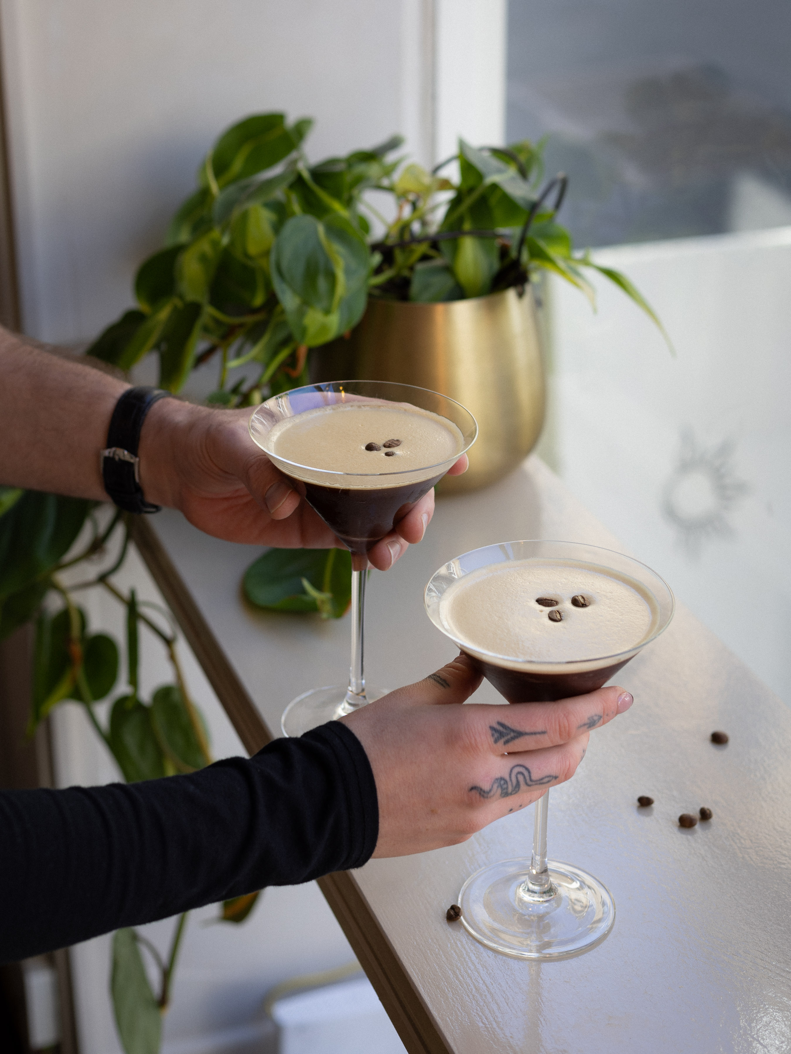 Two espresso martinis topped with coffee beans being handed across a windowsill with a brass-potted houseplant