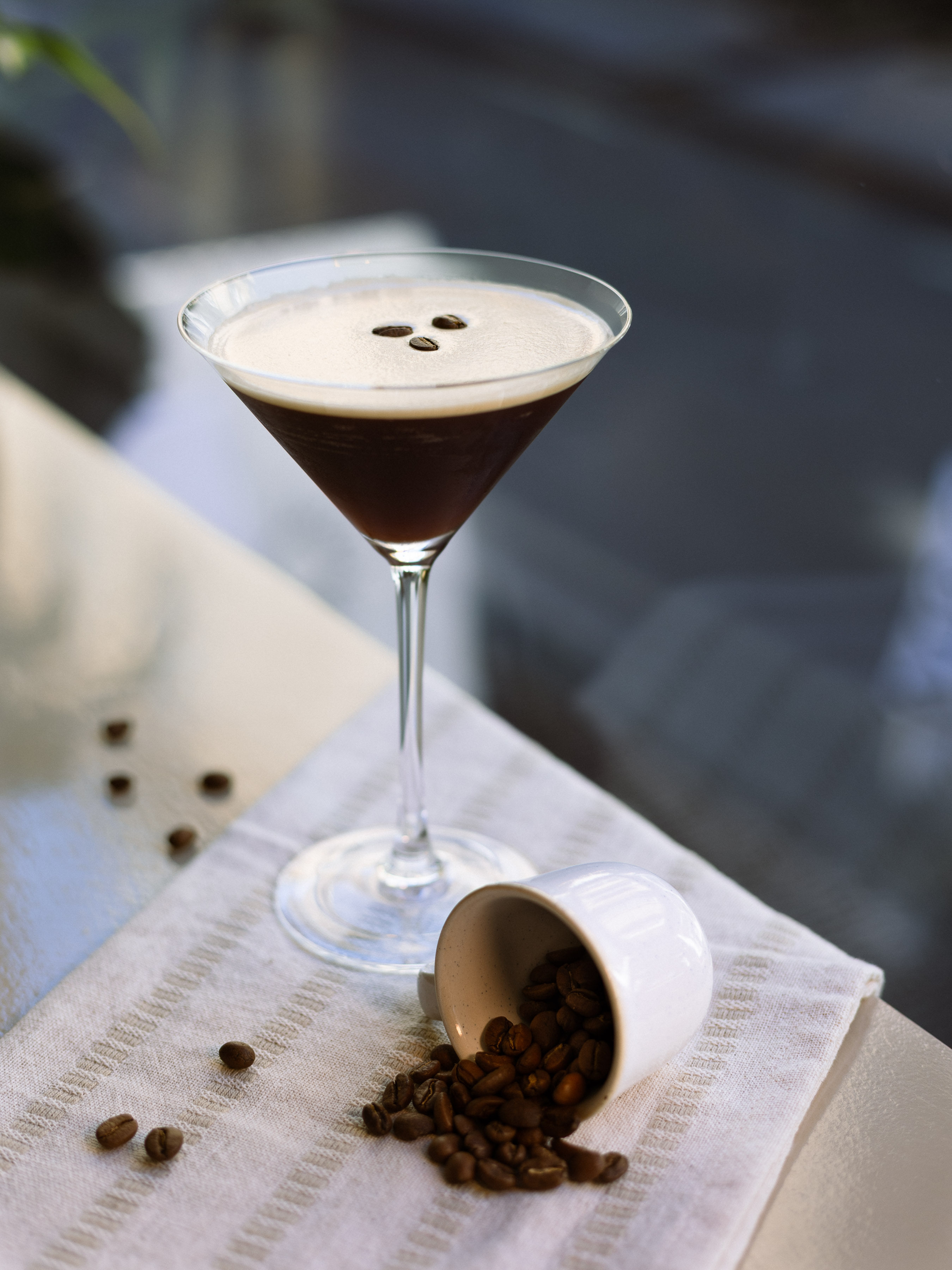 Single espresso martini in a classic glass with coffee beans spilling from a small ceramic cup on a striped linen napkin
