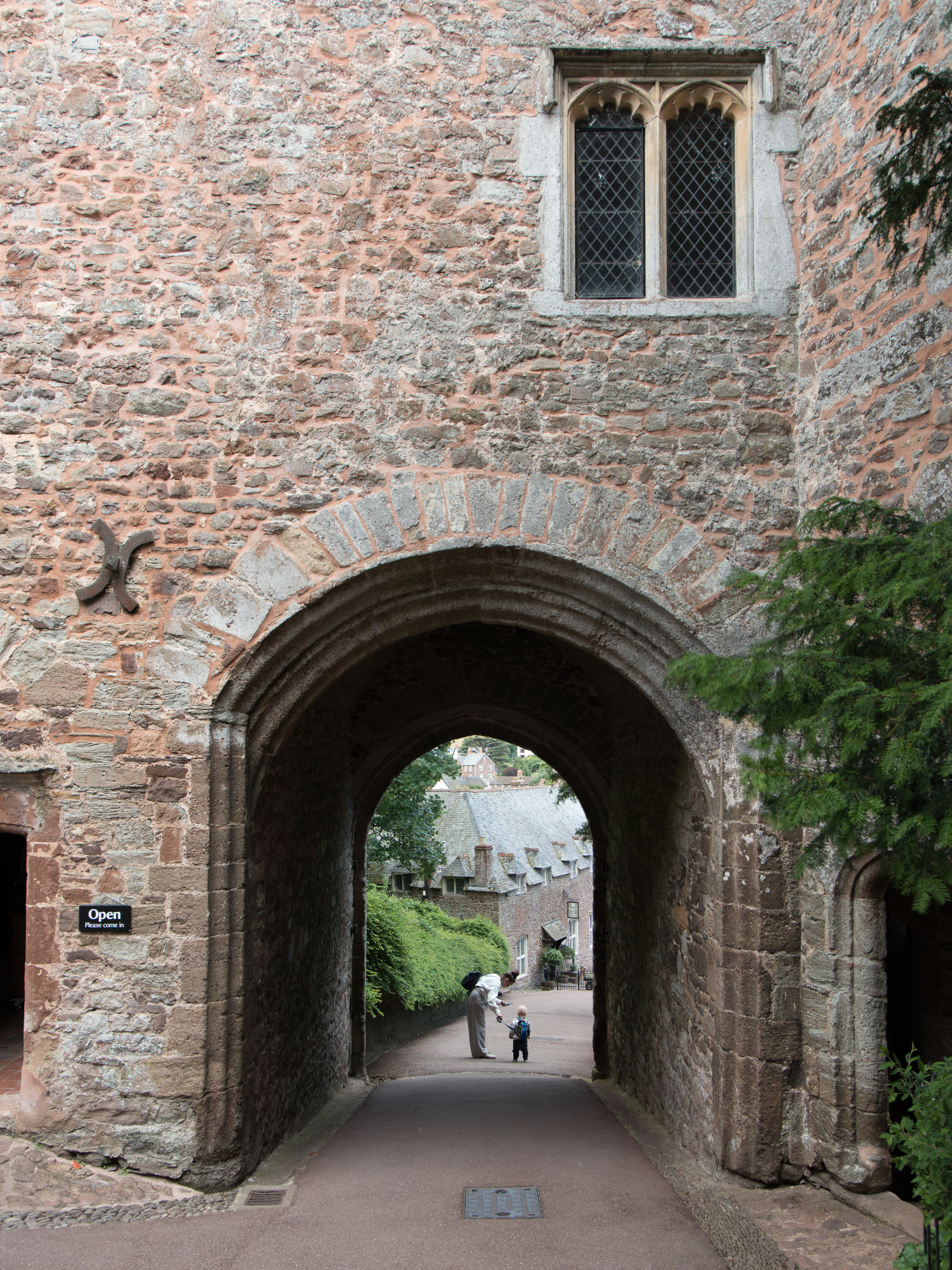 Dunster Castle, worn stone archway in a sandstone castle wall framing a sunlit medieval village street beyond