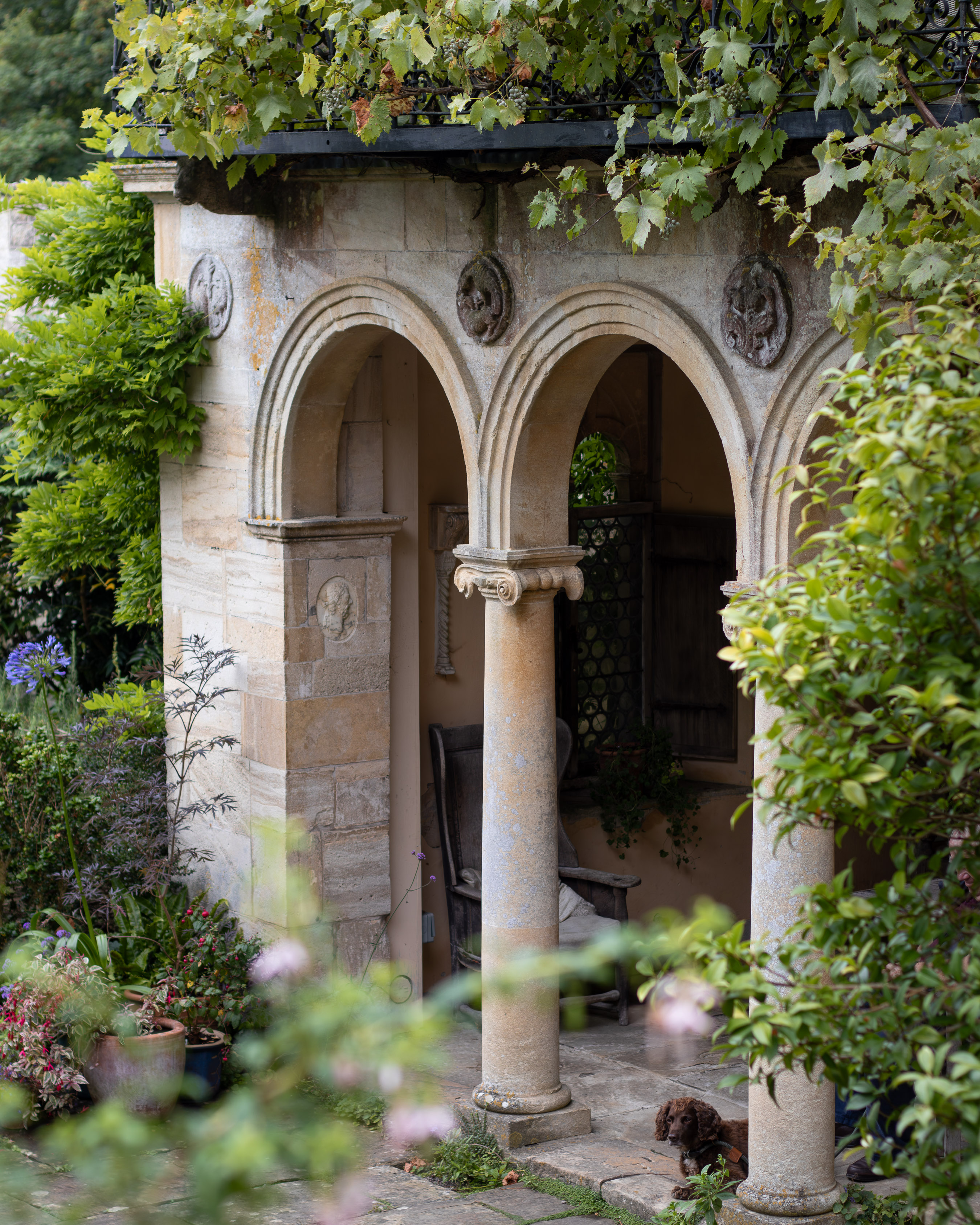 Iford Manor and Gardens, Italianate stone loggia with twin columns and carved roundels, framed by greenery and a spaniel resting in the shade