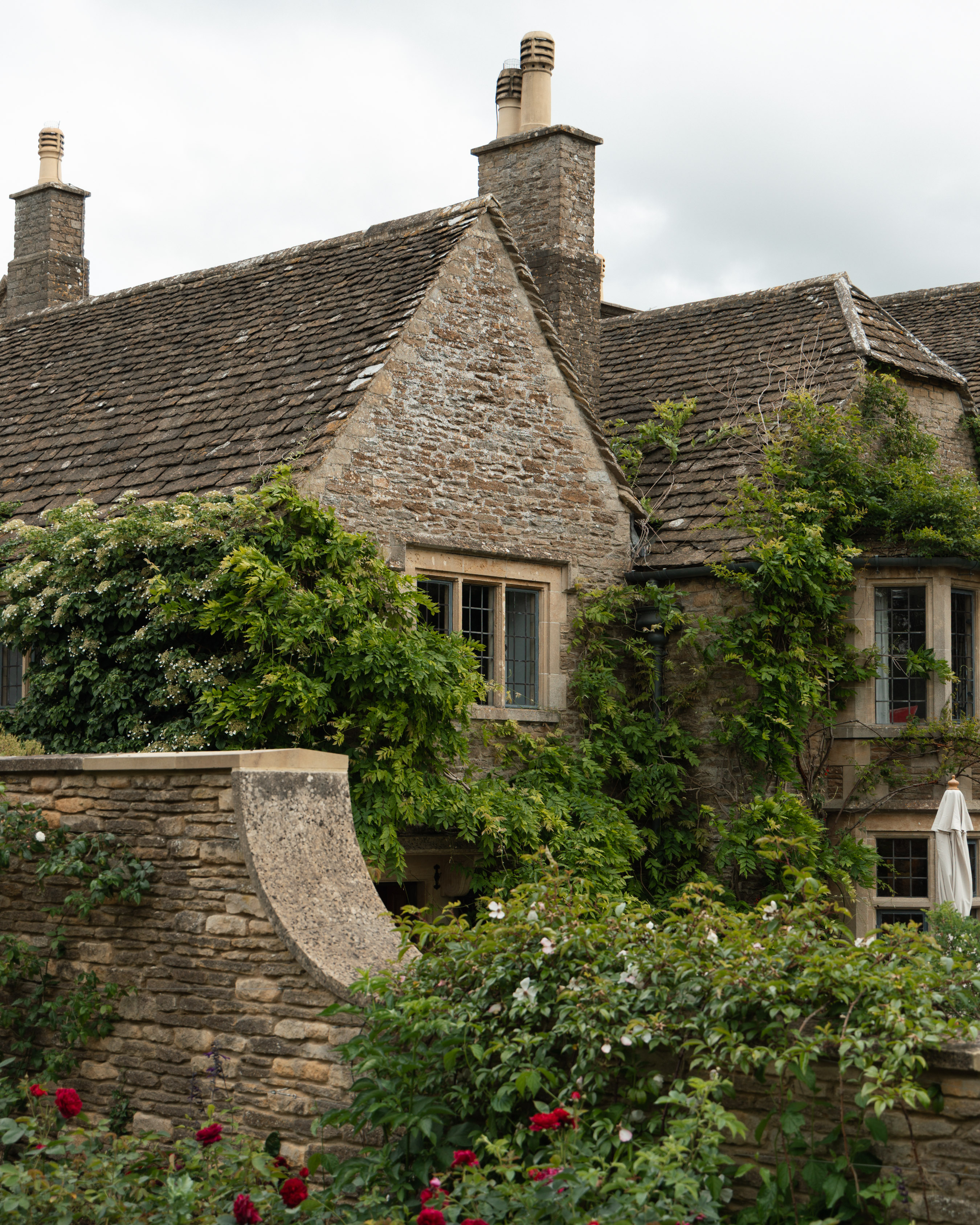 Cotswold-stone cottage with steep tiled roof, climbing wisteria and roses peeking over a drystone wall