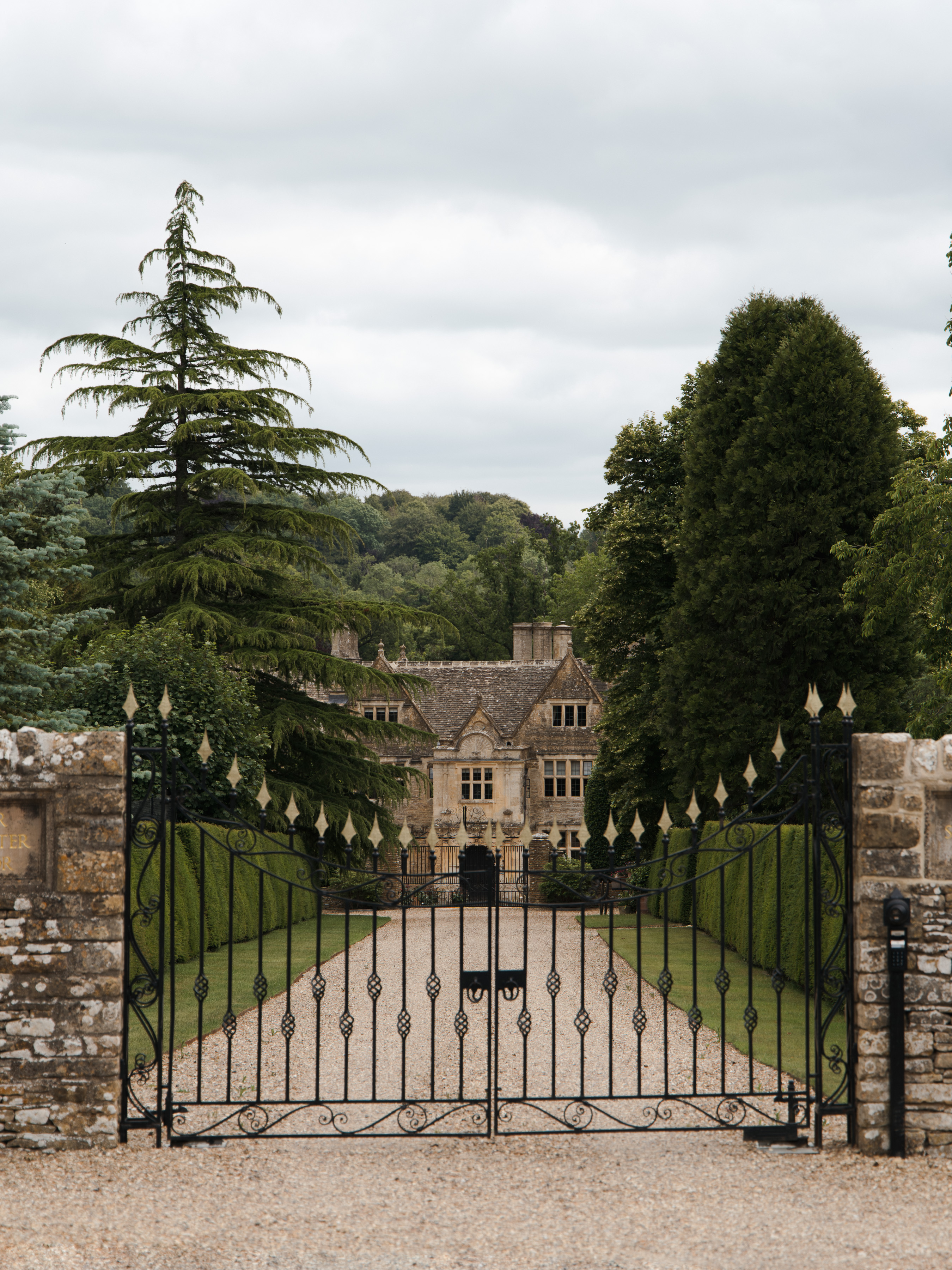 Elizabethan manor house seen through wrought-iron gates flanked by a tall cedar and clipped yews