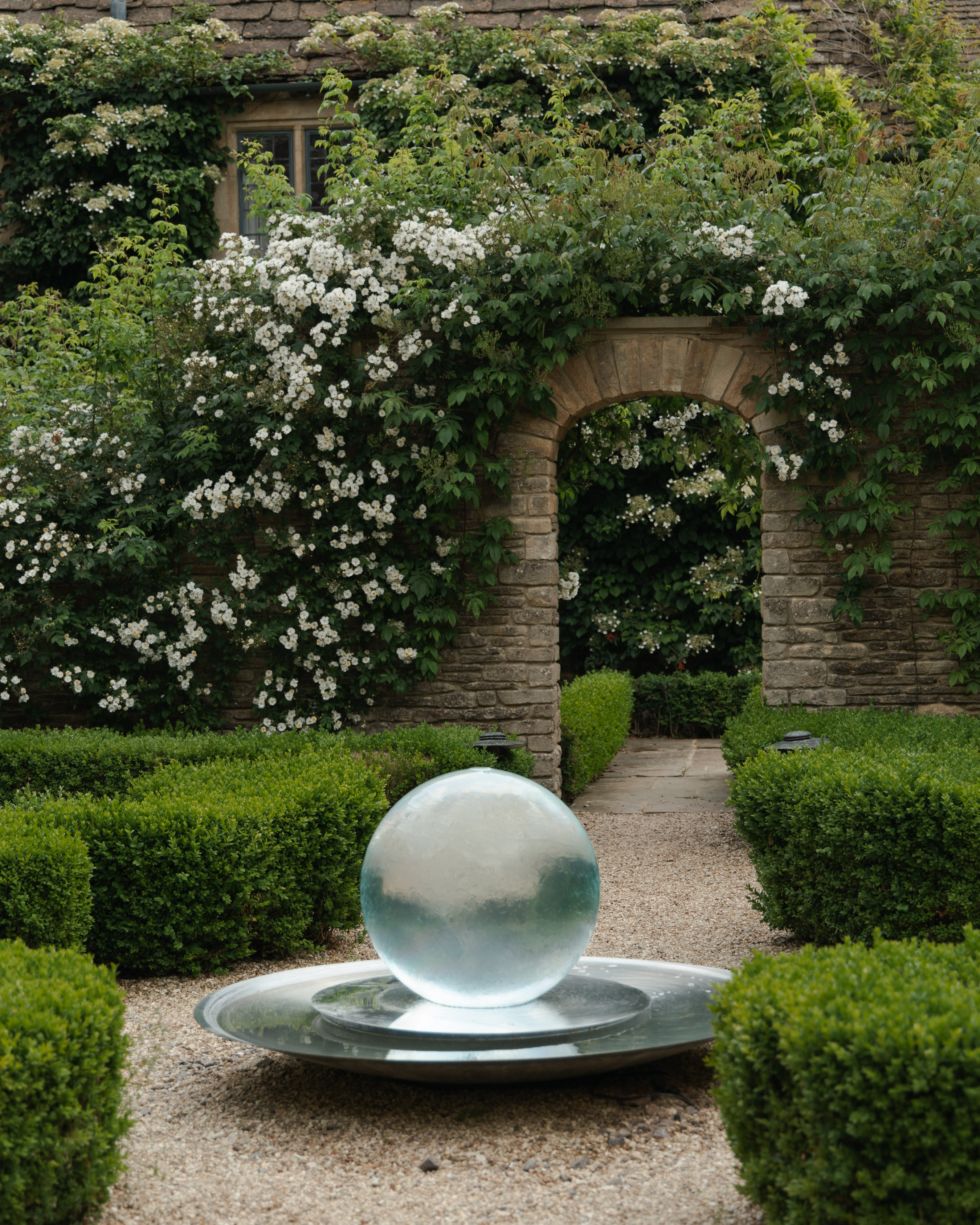 Formal Cotswold parterre garden with clipped box hedges around a glass-globe water feature, framed by a rose-covered stone arch