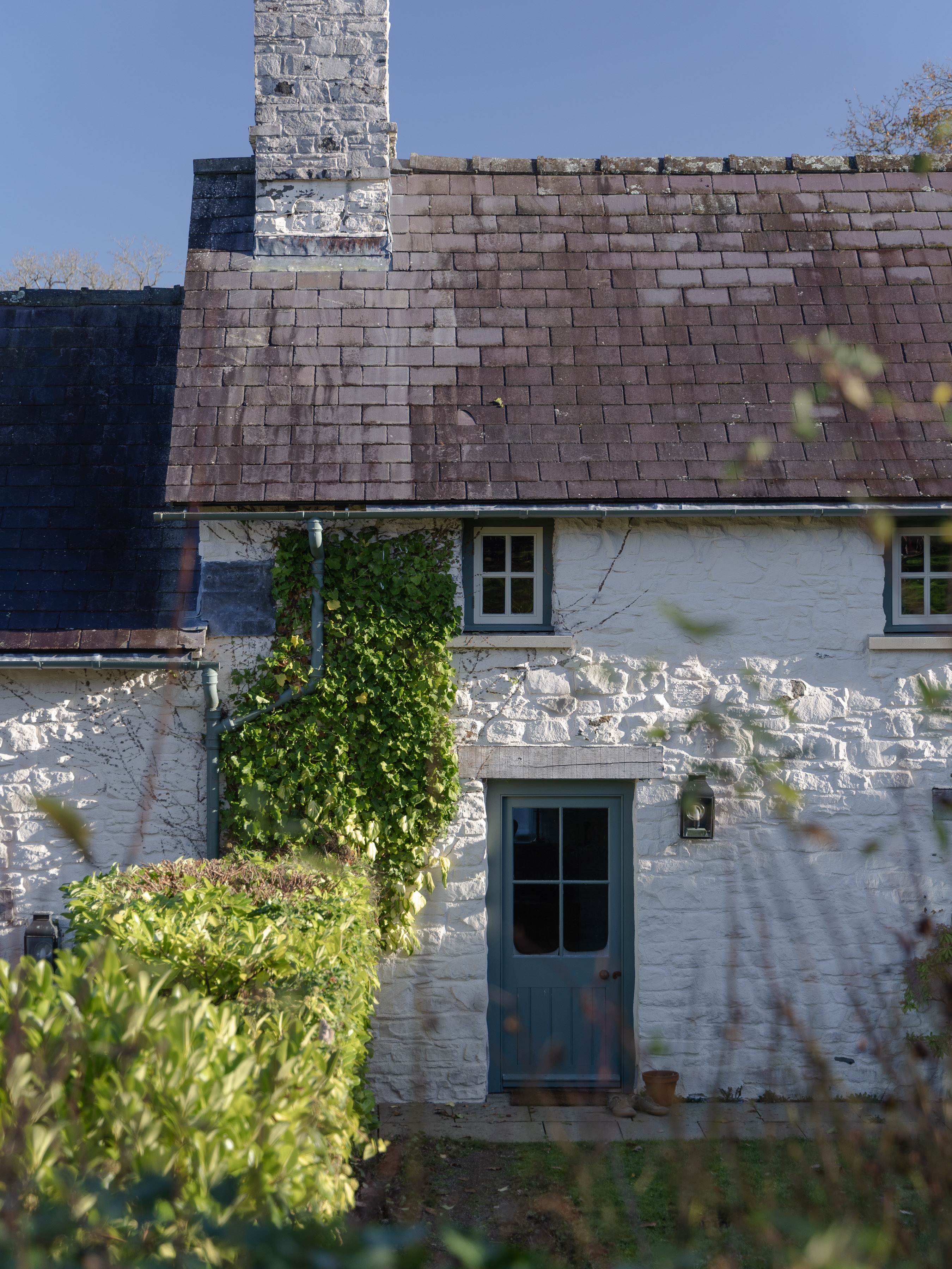 Whitewashed stone Welsh longhouse with slate roof and blue cottage door, framed by autumn hedgerow