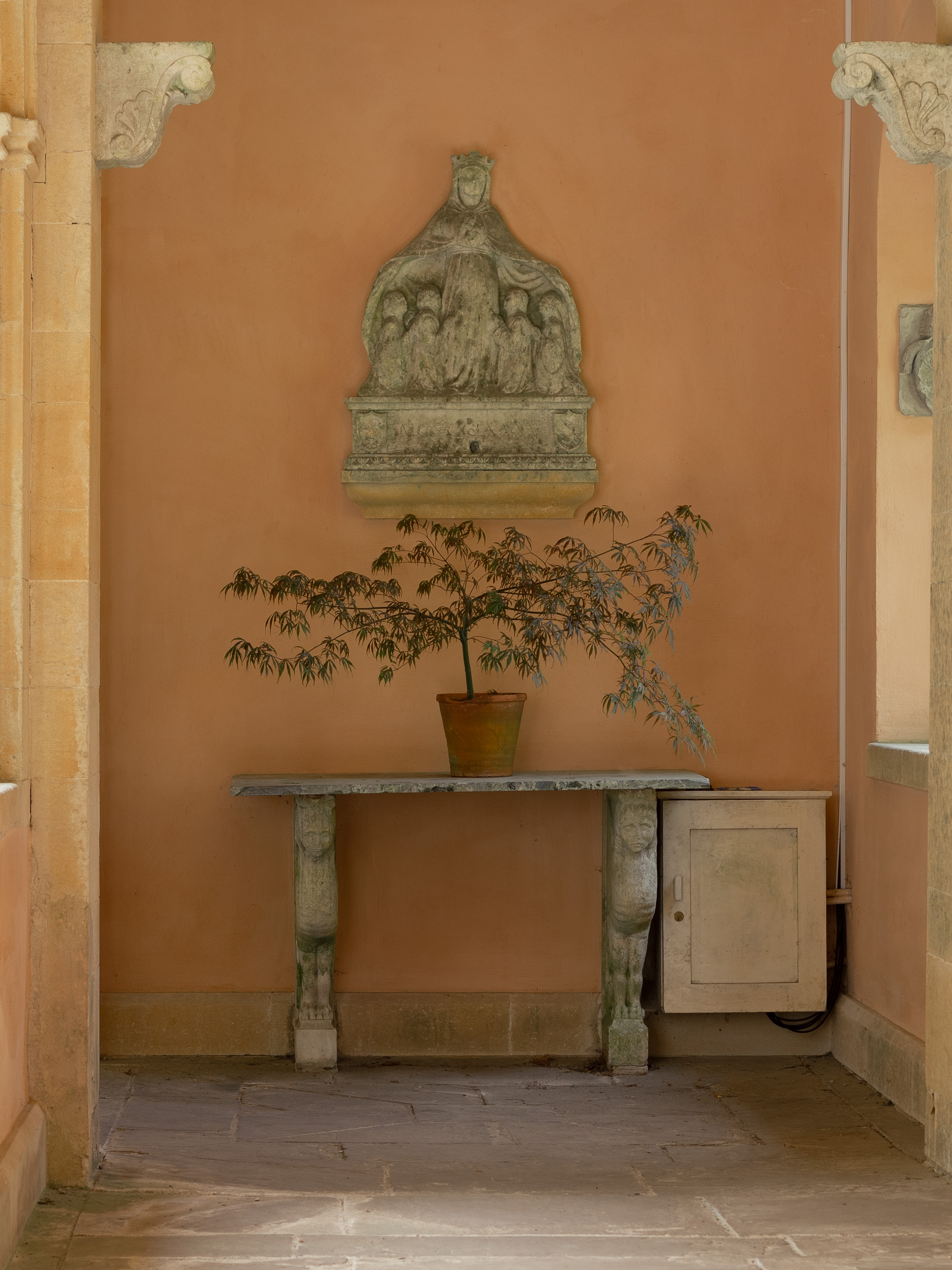 Iford Manor and Gardens, weathered stone relief of a crowned figure above a marble console table on carved supports, with a potted Japanese maple against an apricot cloister wall