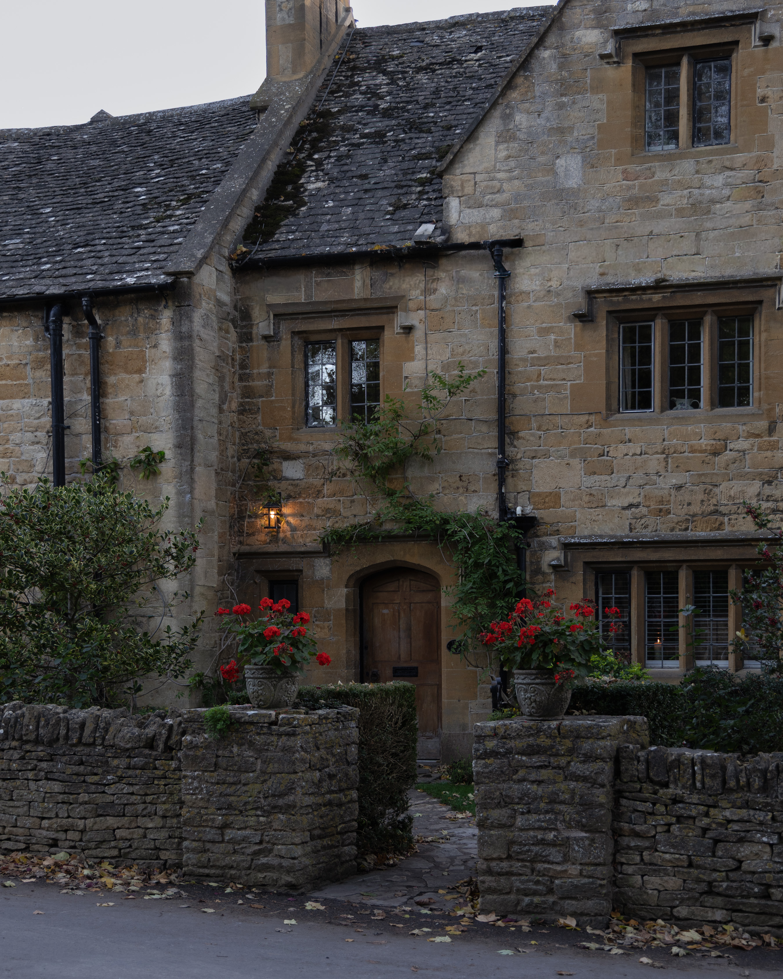 Cotswold-stone house at dusk, scarlet pelargoniums in carved stone urns on a drystone wall, with a lit lantern above the arched oak door and fallen autumn leaves on the lane