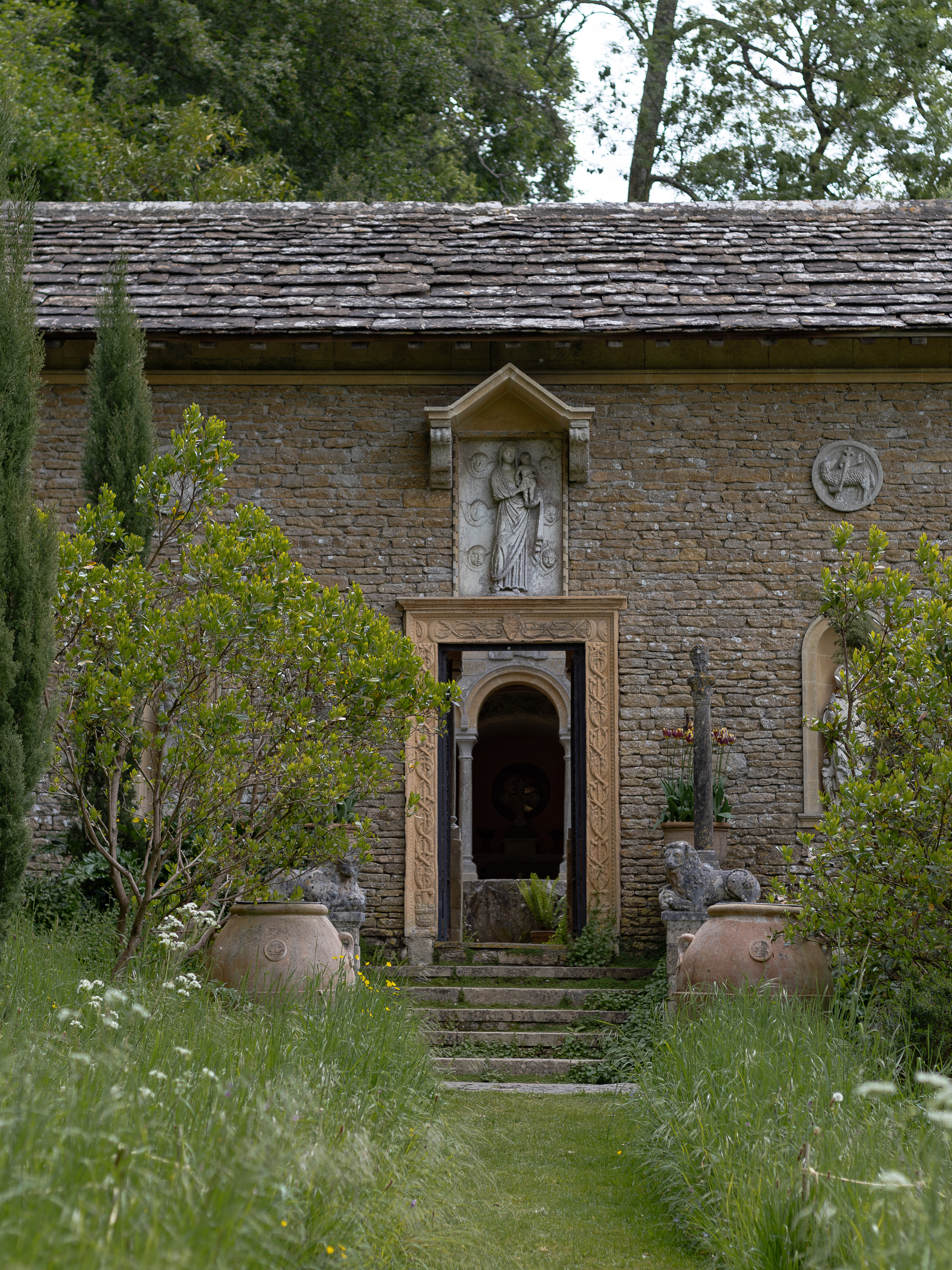 Iford Manor and Gardens, entrance to the Cloisters, a carved Madonna and Child relief set above the ornate stone doorway, flanked by terracotta olive jars, clipped bay and a slender cypress, approached through long meadow grass and cow parsley