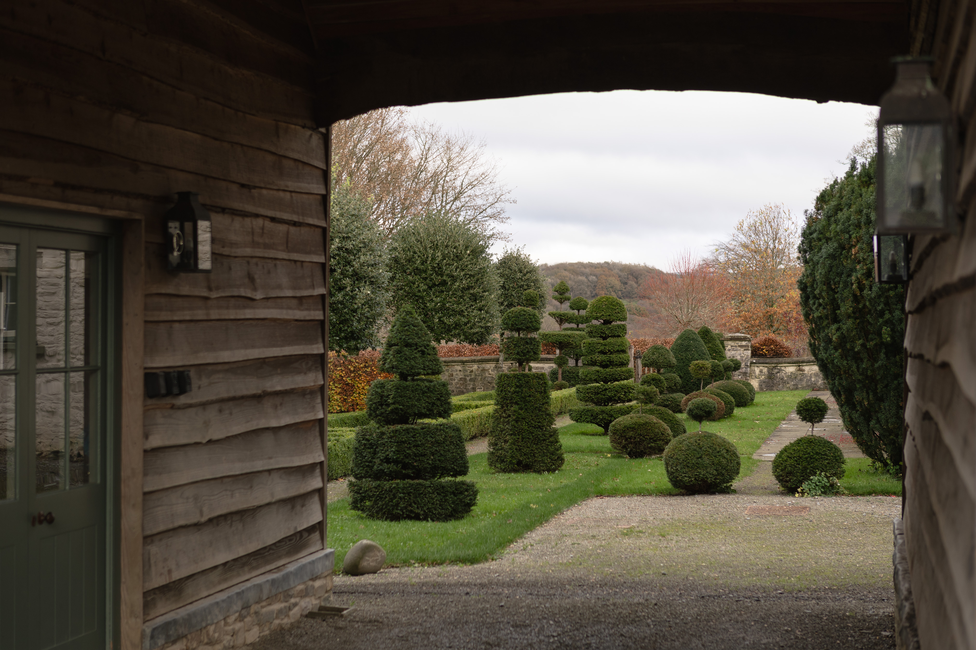 View through an oak-clad barn archway onto a topiary garden of clipped yew cones, spirals and domes, with iron wall lanterns, a green glazed door and an autumn hillside beyond