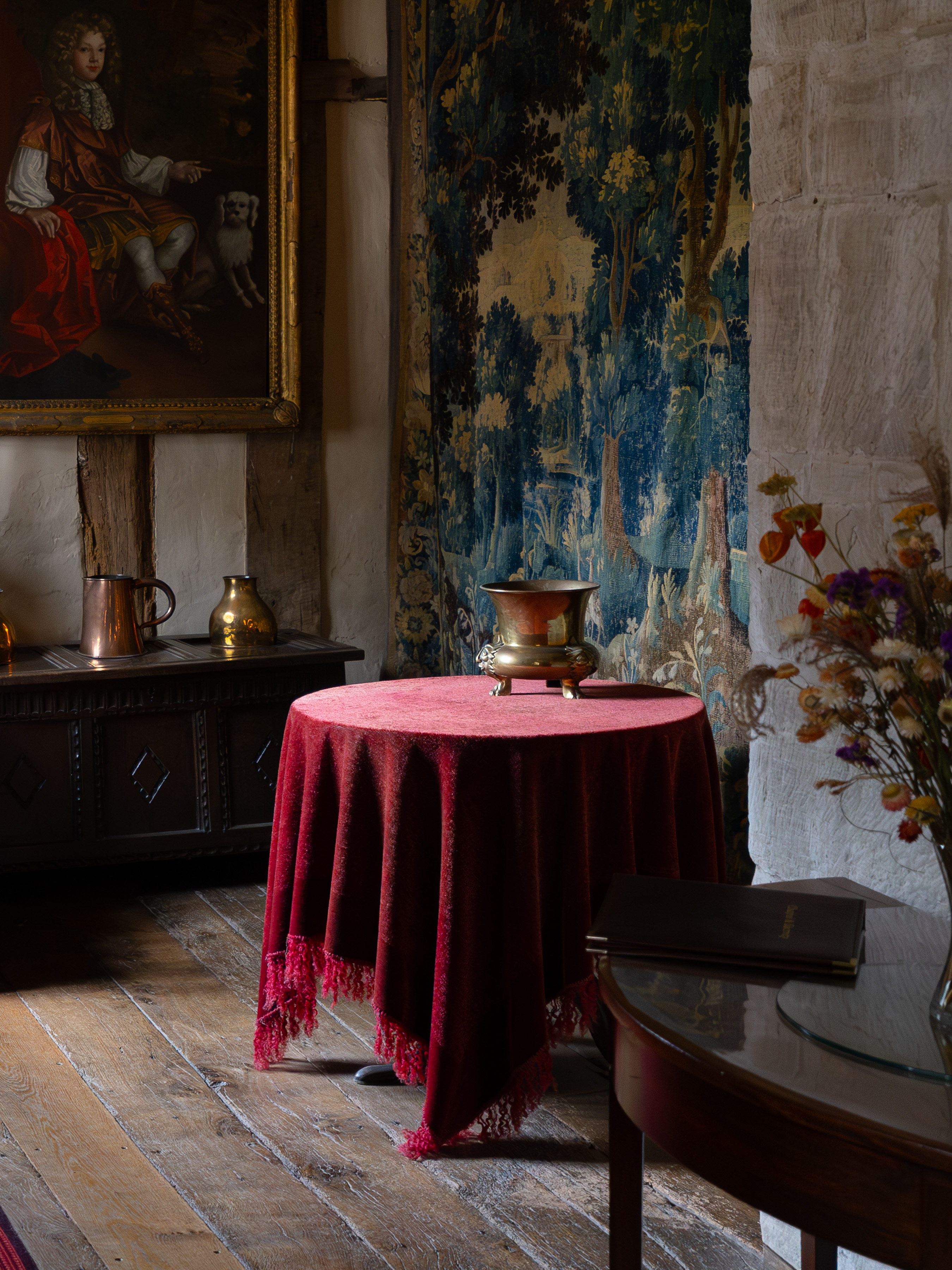 Baddesley Clinton Manor, crimson velvet table beneath antique tapestry, with copper jug, brass urn and dried flowers in a historic stone-walled room