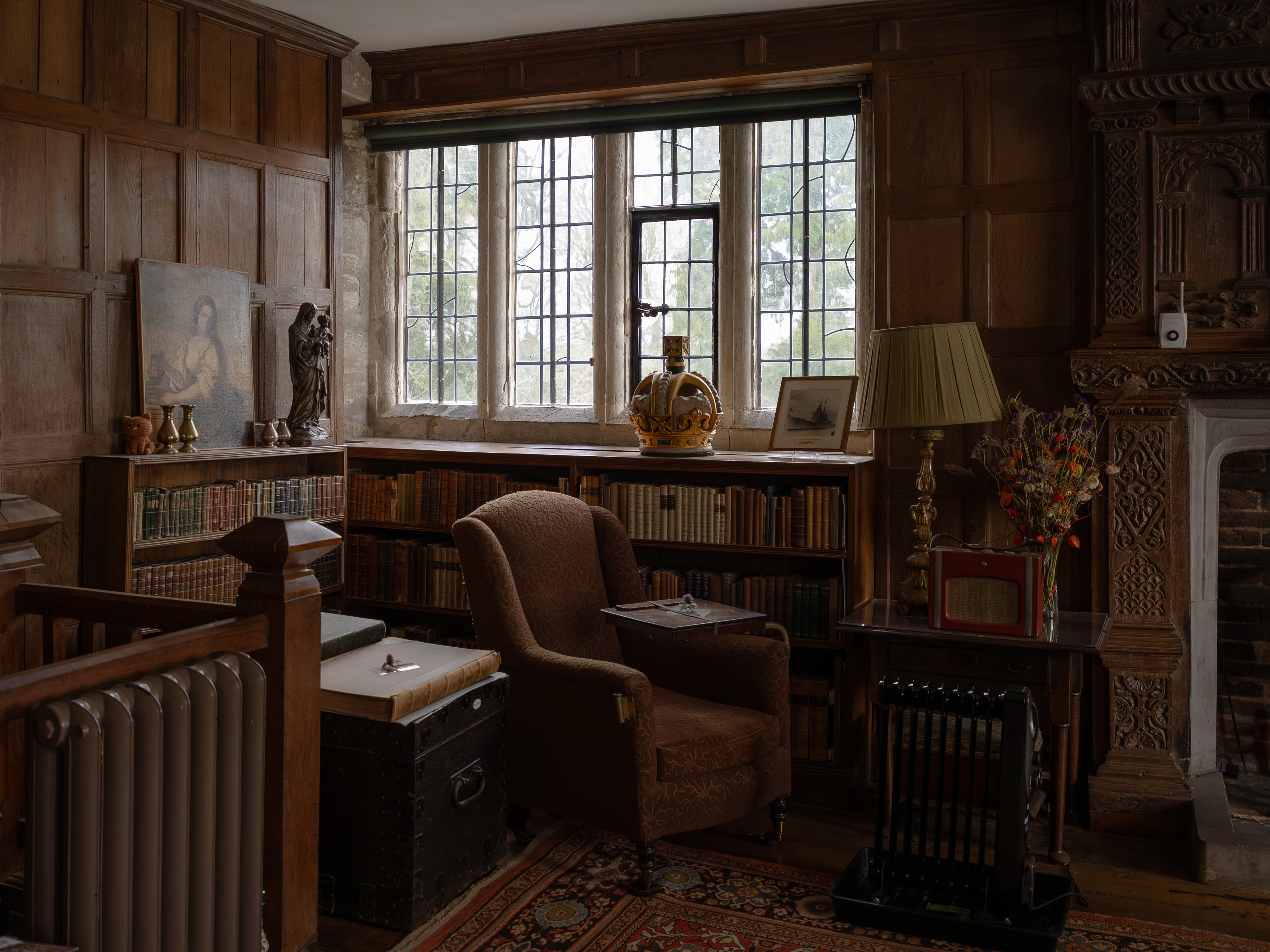 Baddesley Clinton Manor, wood-panelled library with leaded windows, brown armchair, bookcases of antique volumes and a ceremonial crown on the windowsill