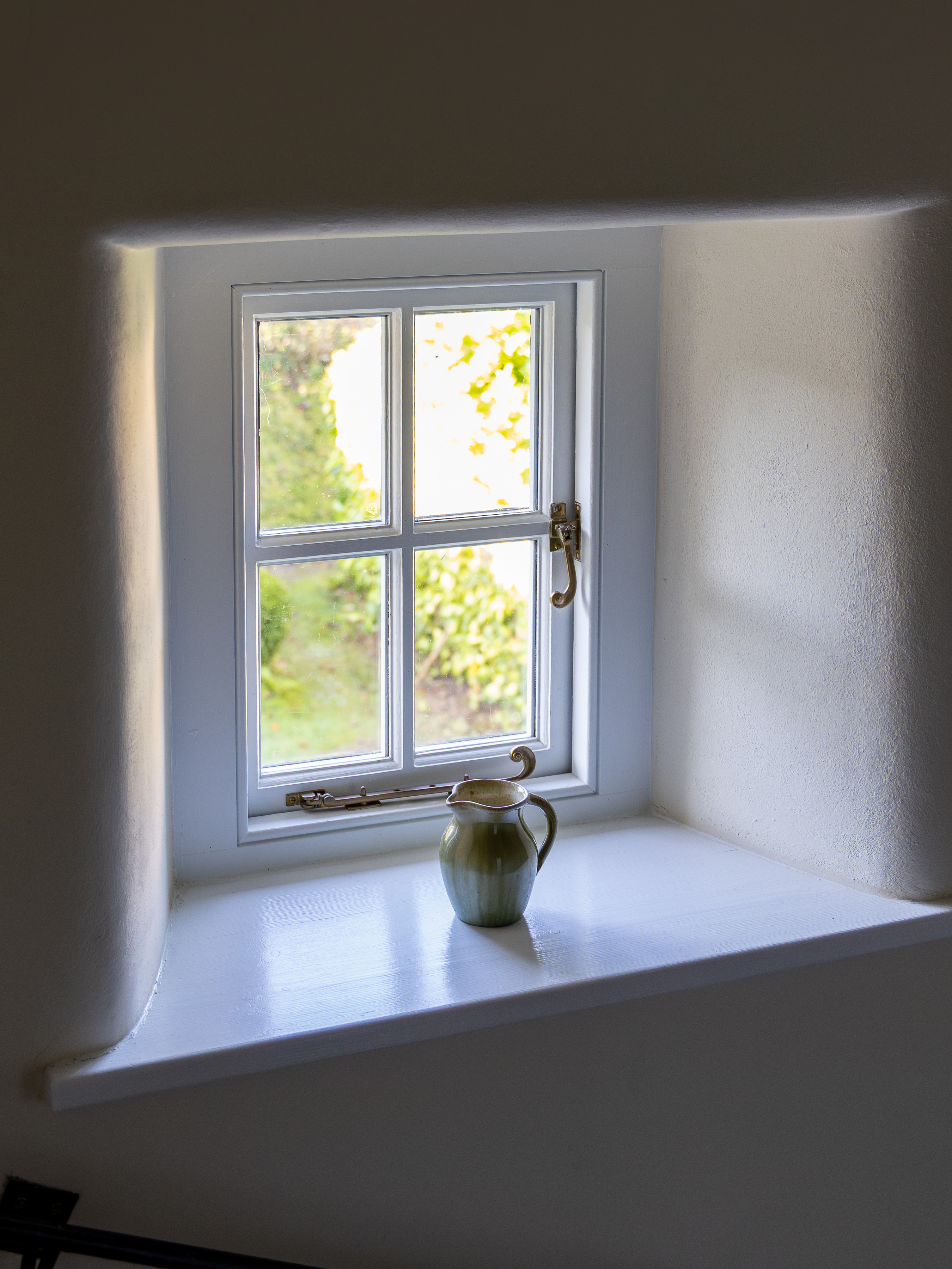 Small white cottage window with a green ceramic jug on the painted sill, soft daylight falling onto a thick render-walled reveal