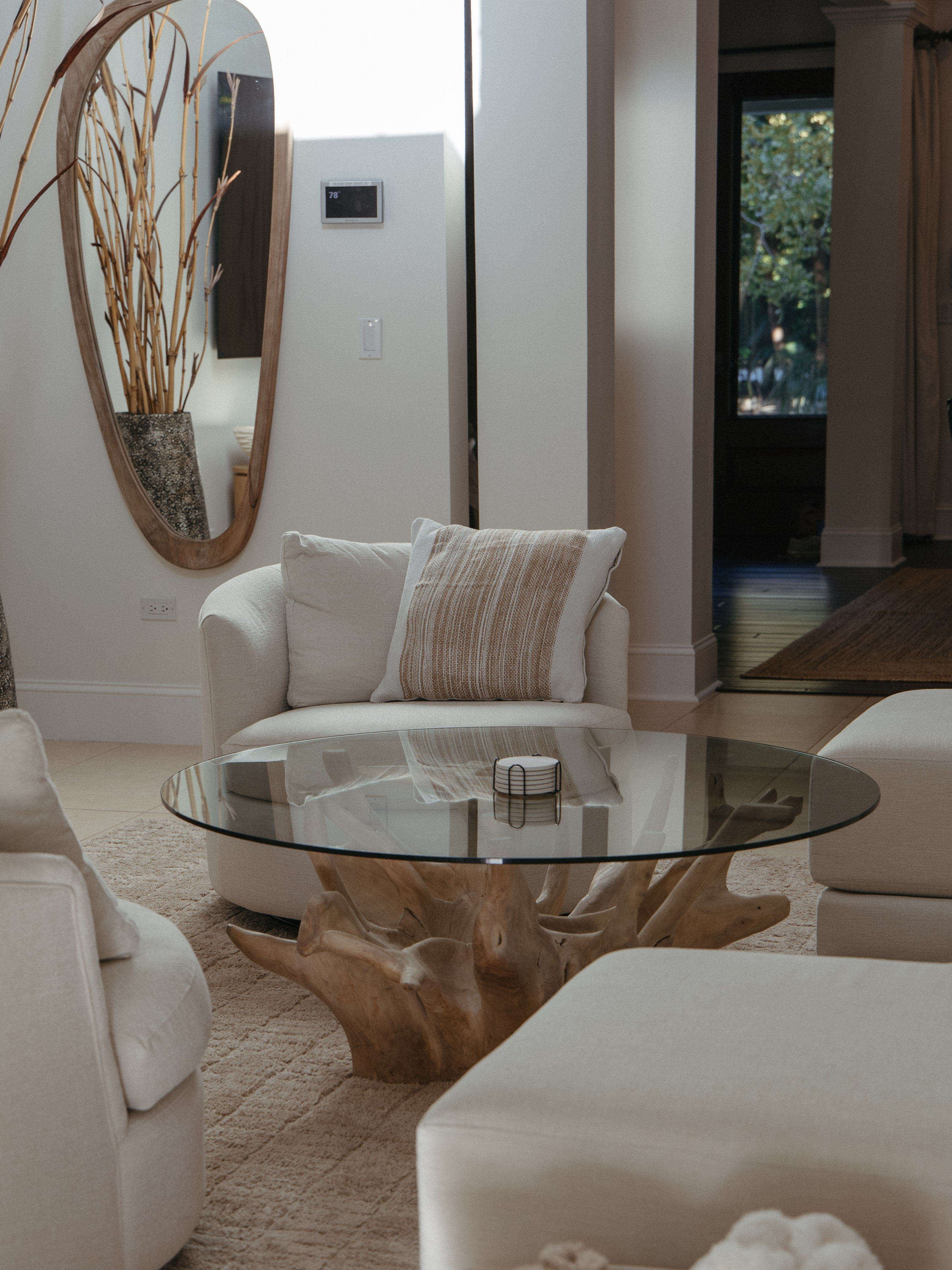 Cream swivel chair and round glass coffee table on a driftwood base, neutral living room with organic mirror and dried grasses