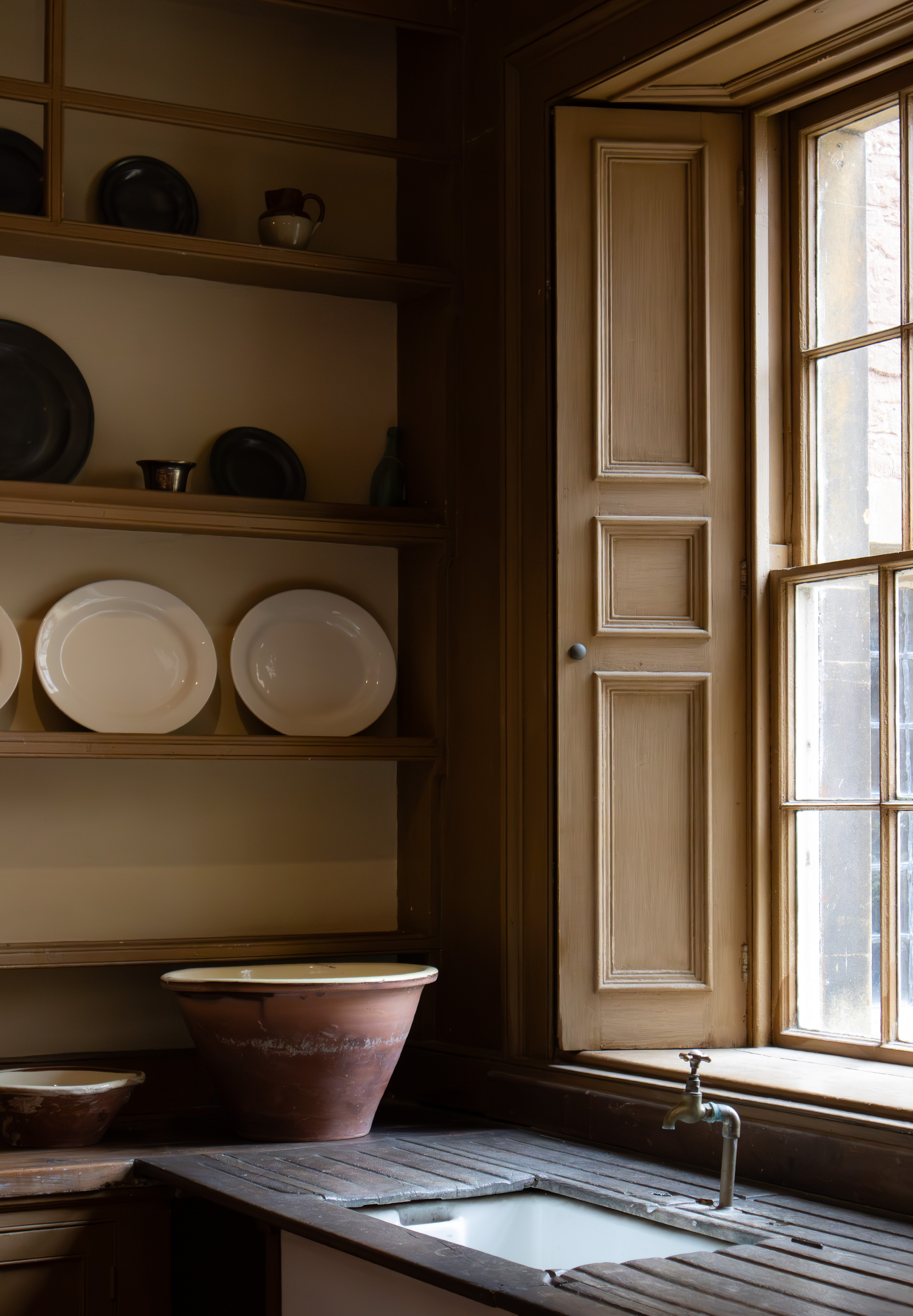 Dyrham Park, quiet stone pantry corner with butler sink, brass tap, terracotta bowls and shelves of pewter and creamware