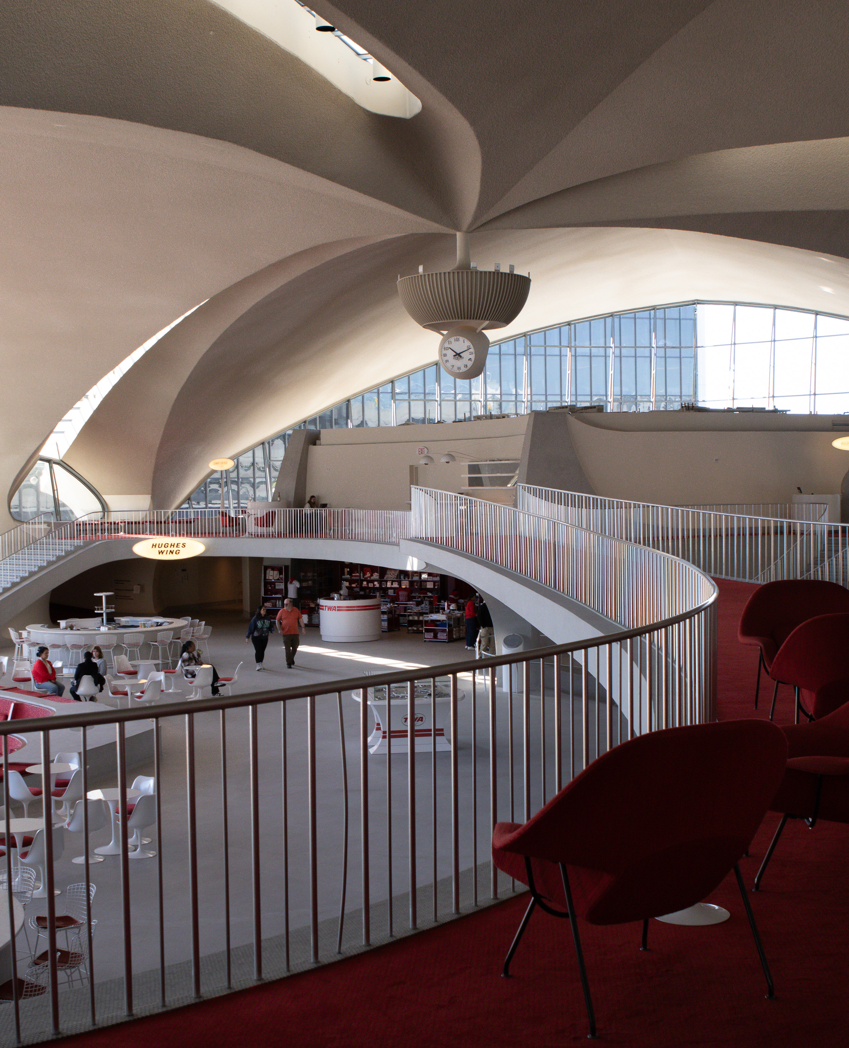 Upper concourse of a mid-century terminal with red carpet, womb chairs and a sculptural clock beneath sweeping concrete arches