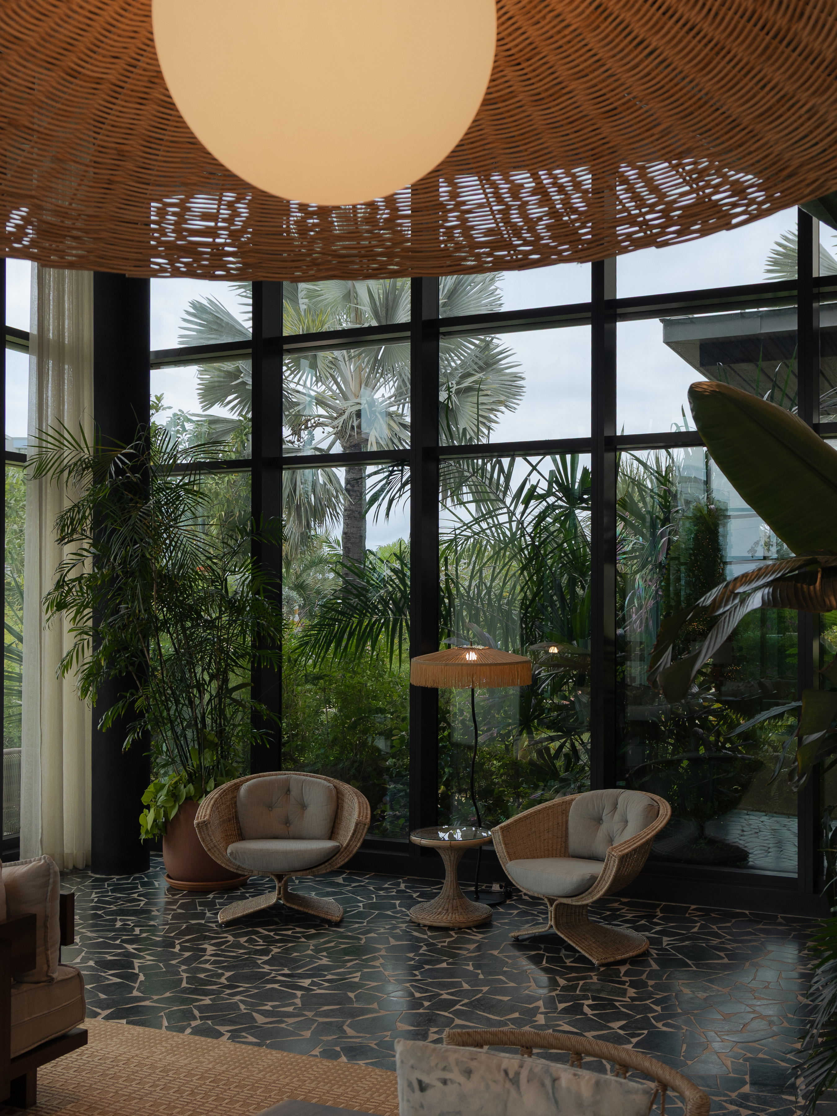 Tropical hotel lounge with woven rattan pendant light, sculptural cane chairs and palm-filled garden beyond glass walls