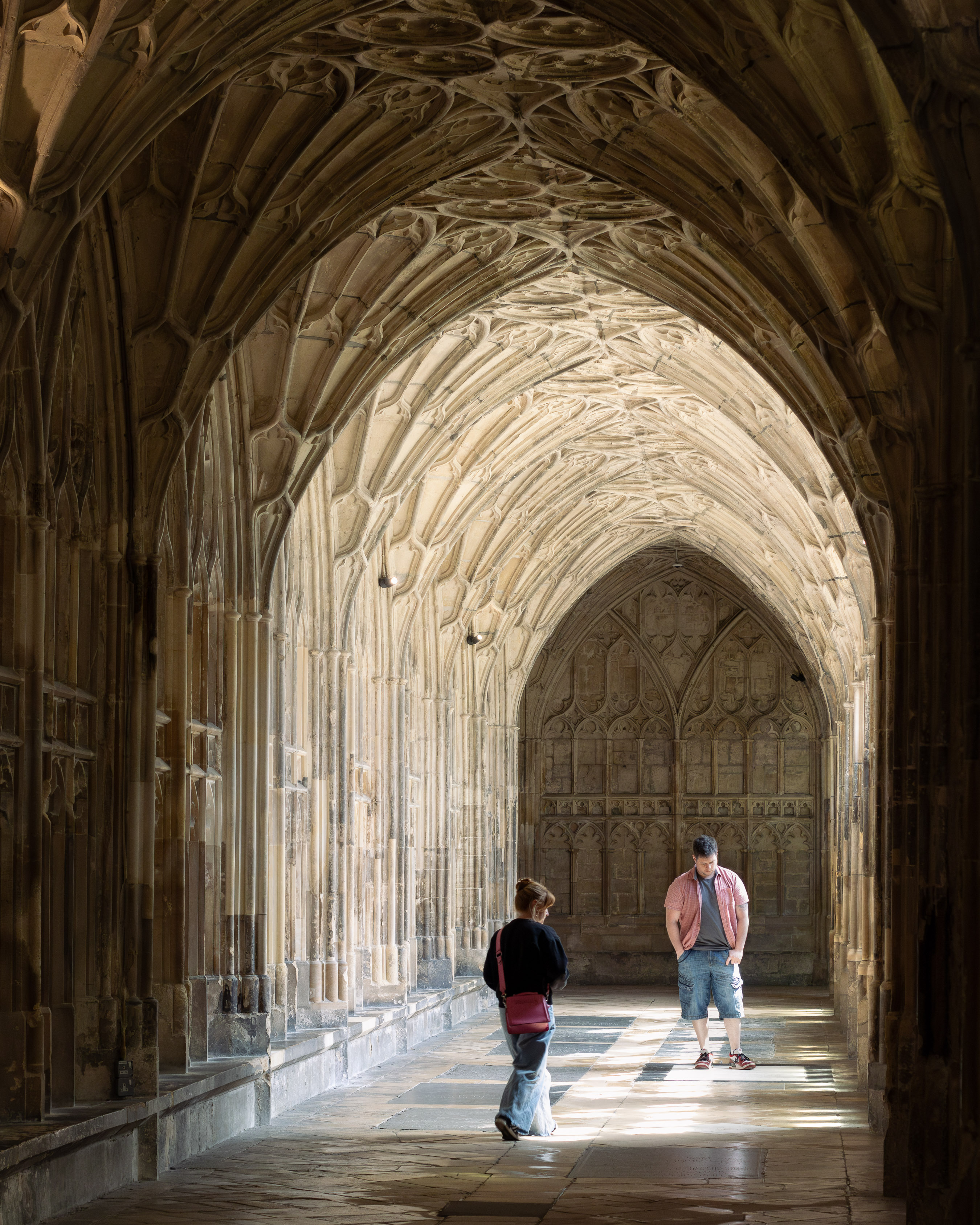 Gloucester Cathedral, two visitors pause in the dappled light of a Gothic cloister with intricate fan-vaulted ceiling