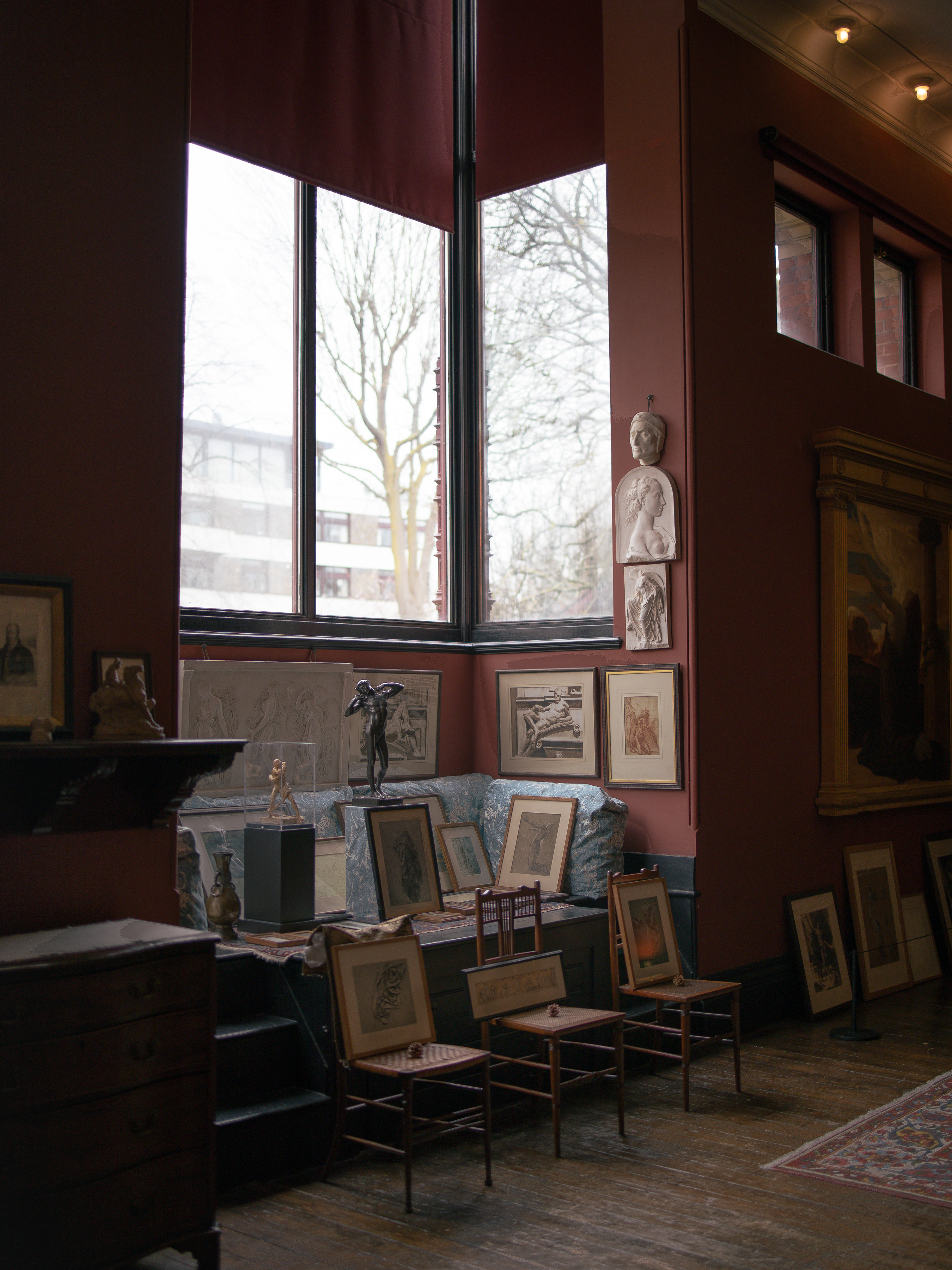 Leighton House, artist's studio with deep oxblood walls, plaster casts and a bronze figure, framed life drawings propped along a row of bentwood chairs beneath a tall studio window