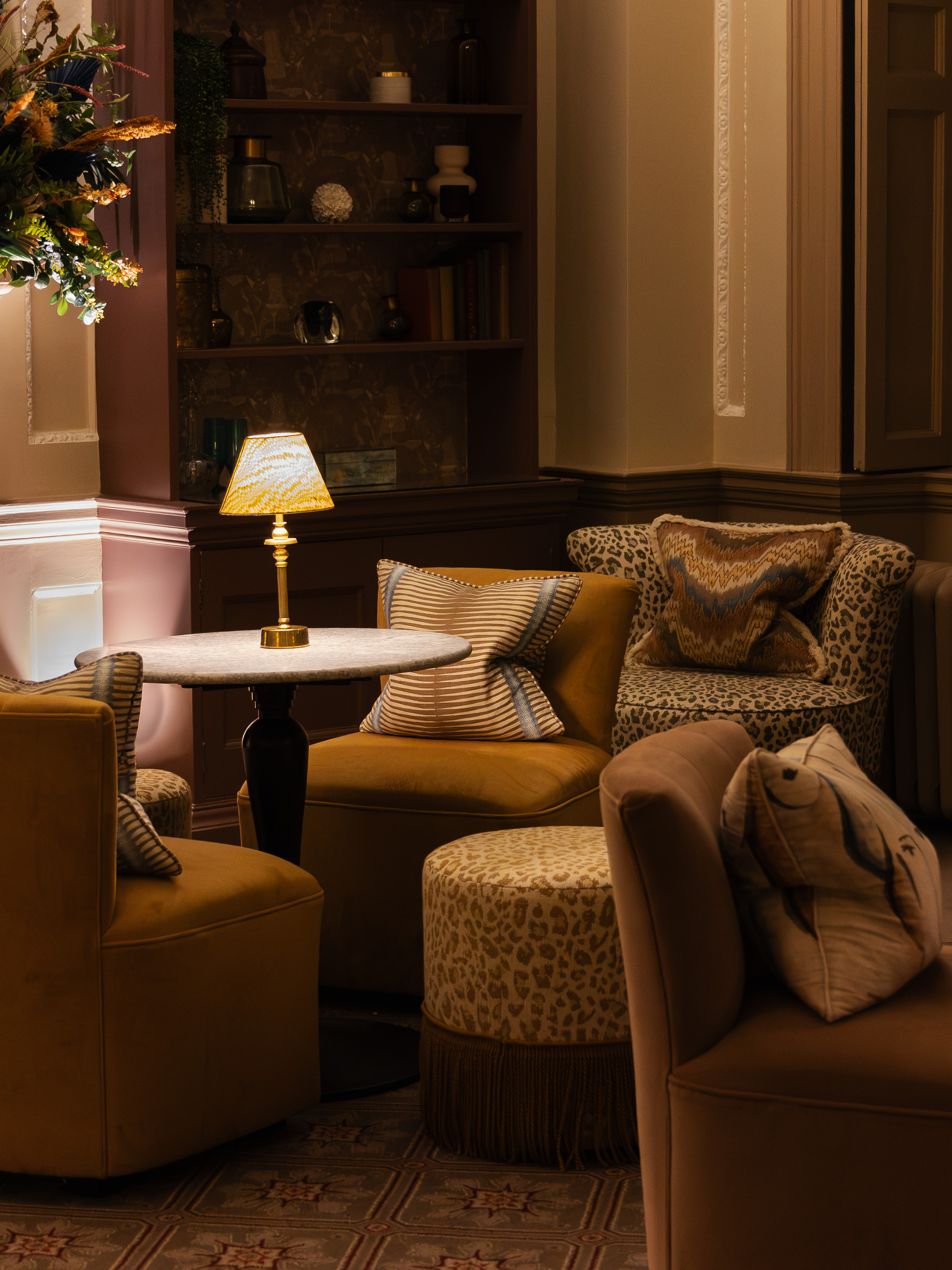 The Royal Crescent Hotel, lamp-lit lounge with mustard velvet tub chairs, a leopard-print armchair and fringed ottoman, striped and ikat cushions, and a brass lamp on a marble pedestal table against dusky pink panelling