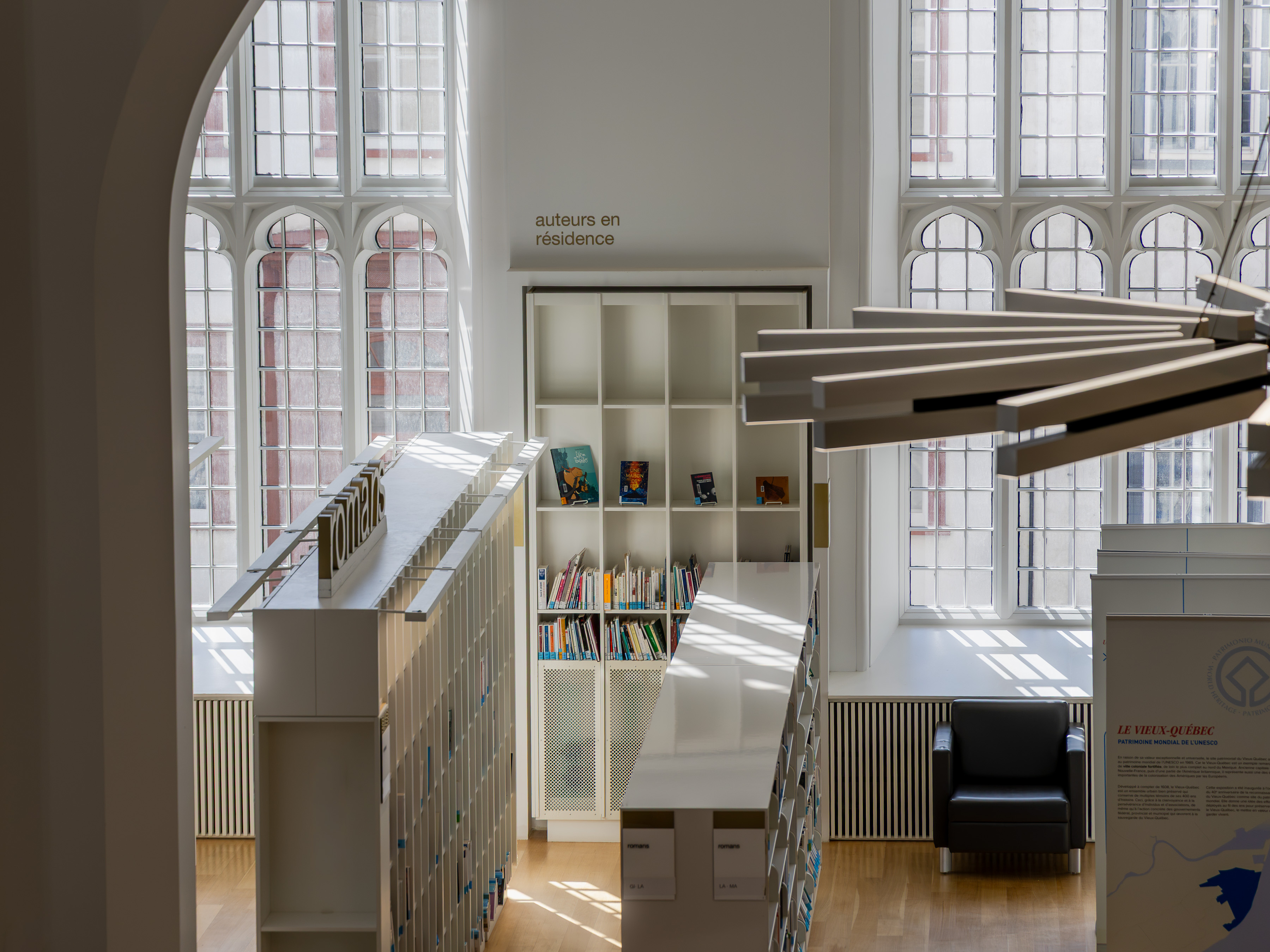 Maison de la littérature, library inside a converted neo-Gothic church, white shelving and an 'auteurs en résidence' alcove set against soaring traceried windows, with sunlight falling across pale oak floors