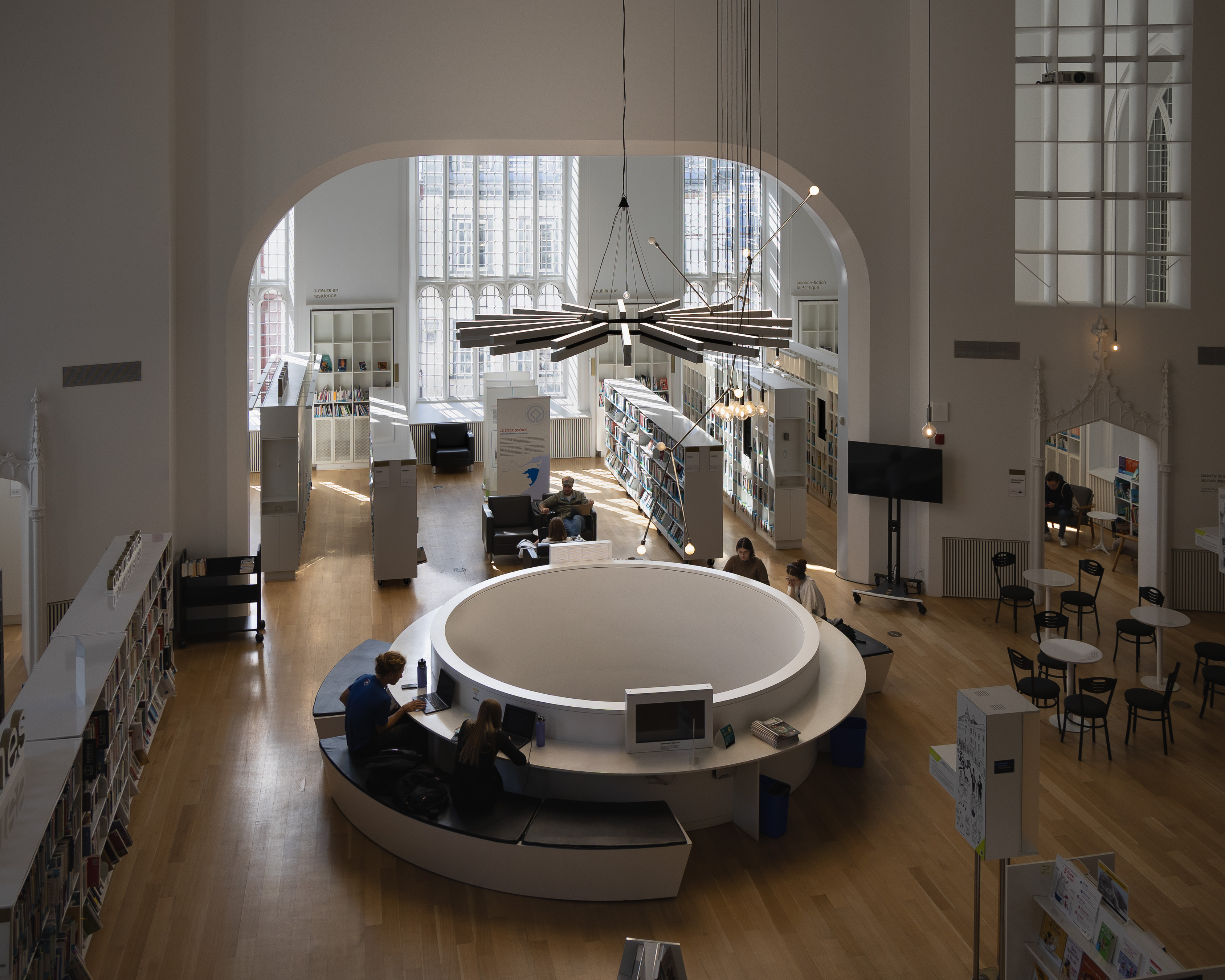 Maison de la littérature, former church remade as a library, with a circular white study platform and radial pendant light beneath tall Gothic windows, readers at work along the book stacks
